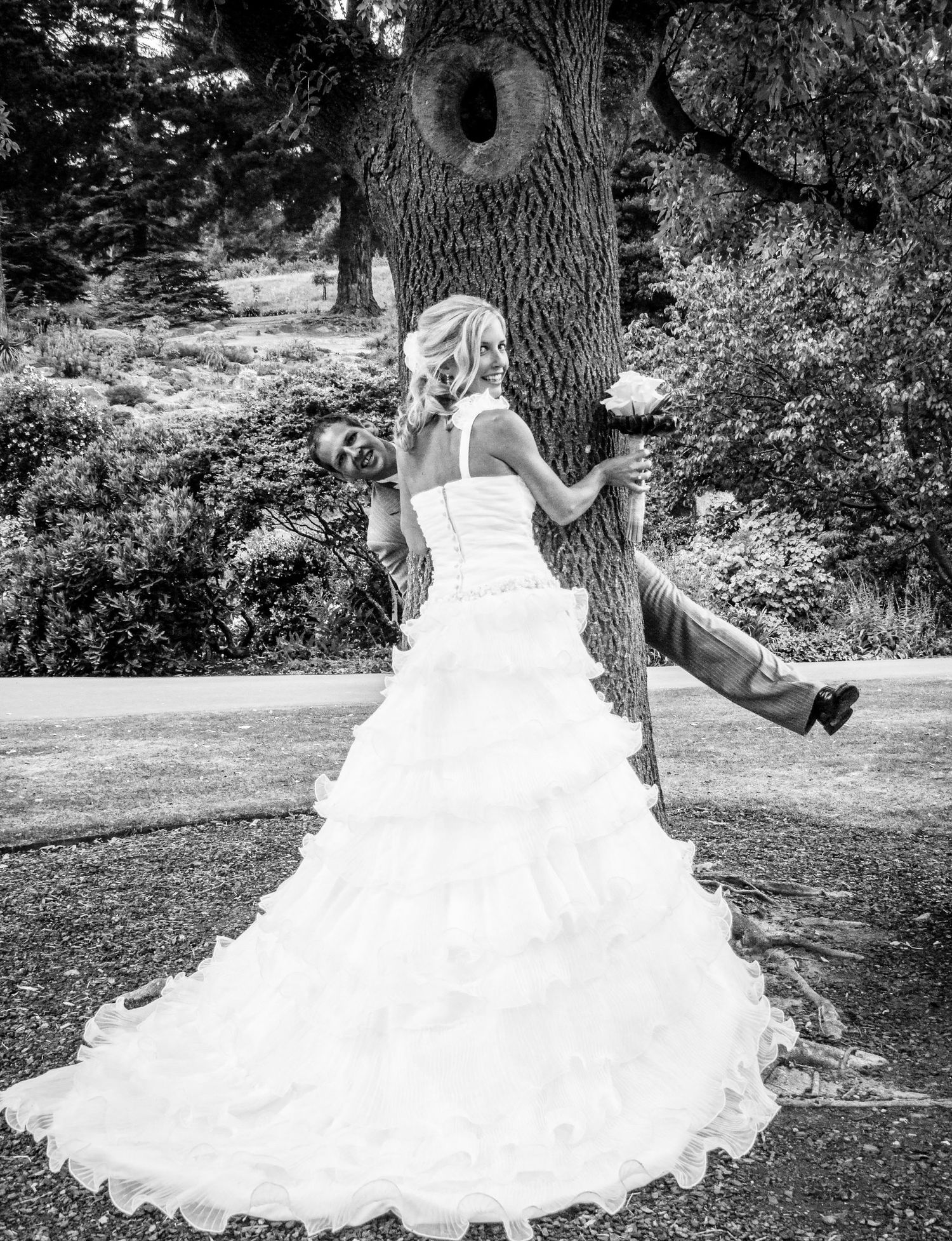 Photo de mariage en noir et blanc, le marié s'amusant à se cacher derrière un grand arbre près de sa femme, par Jérémie Botte.
