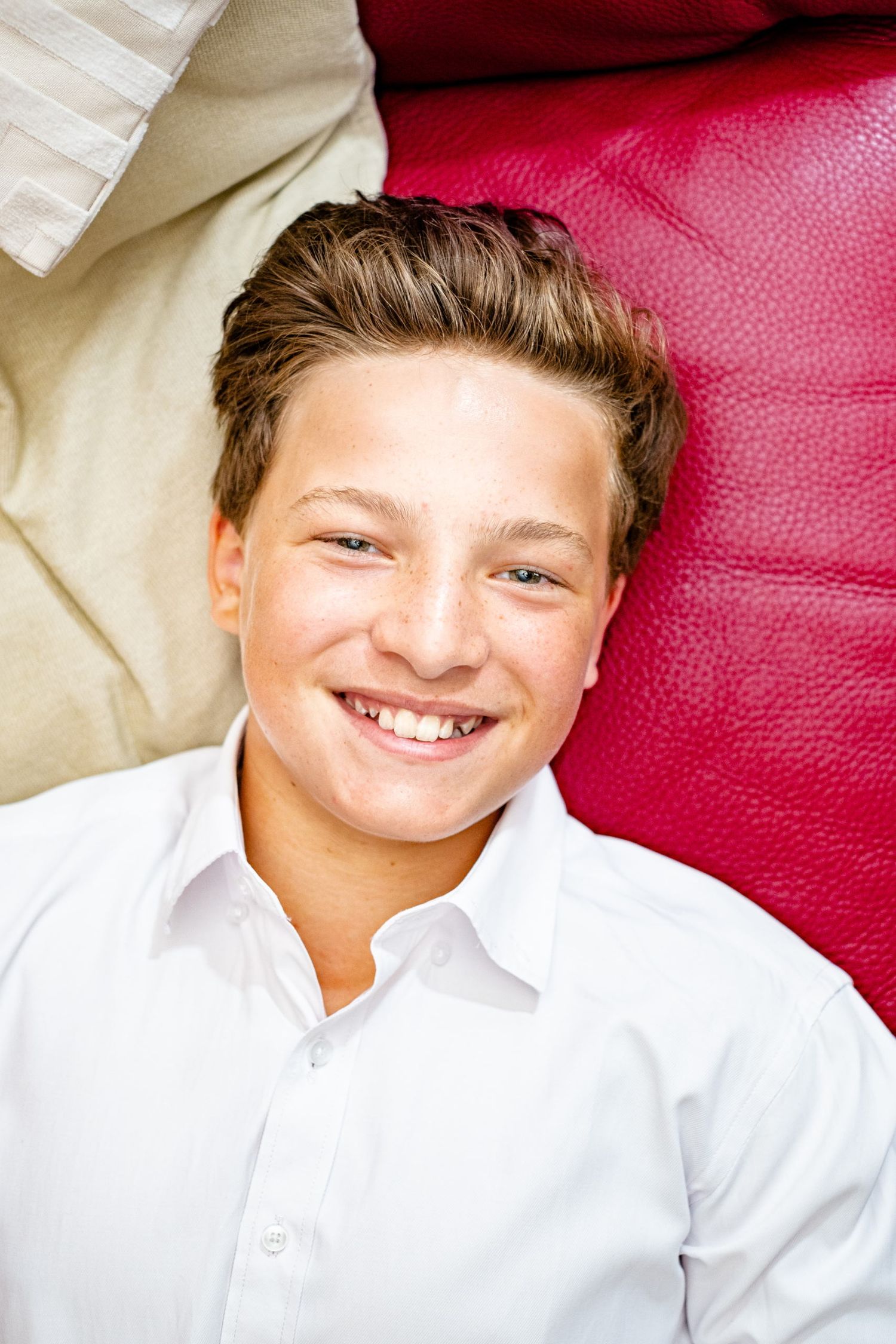 Portrait d'un jeune garçon souriant, allongé de biais entre un oreiller clair et un canapé rouge par Jérémie Botte.