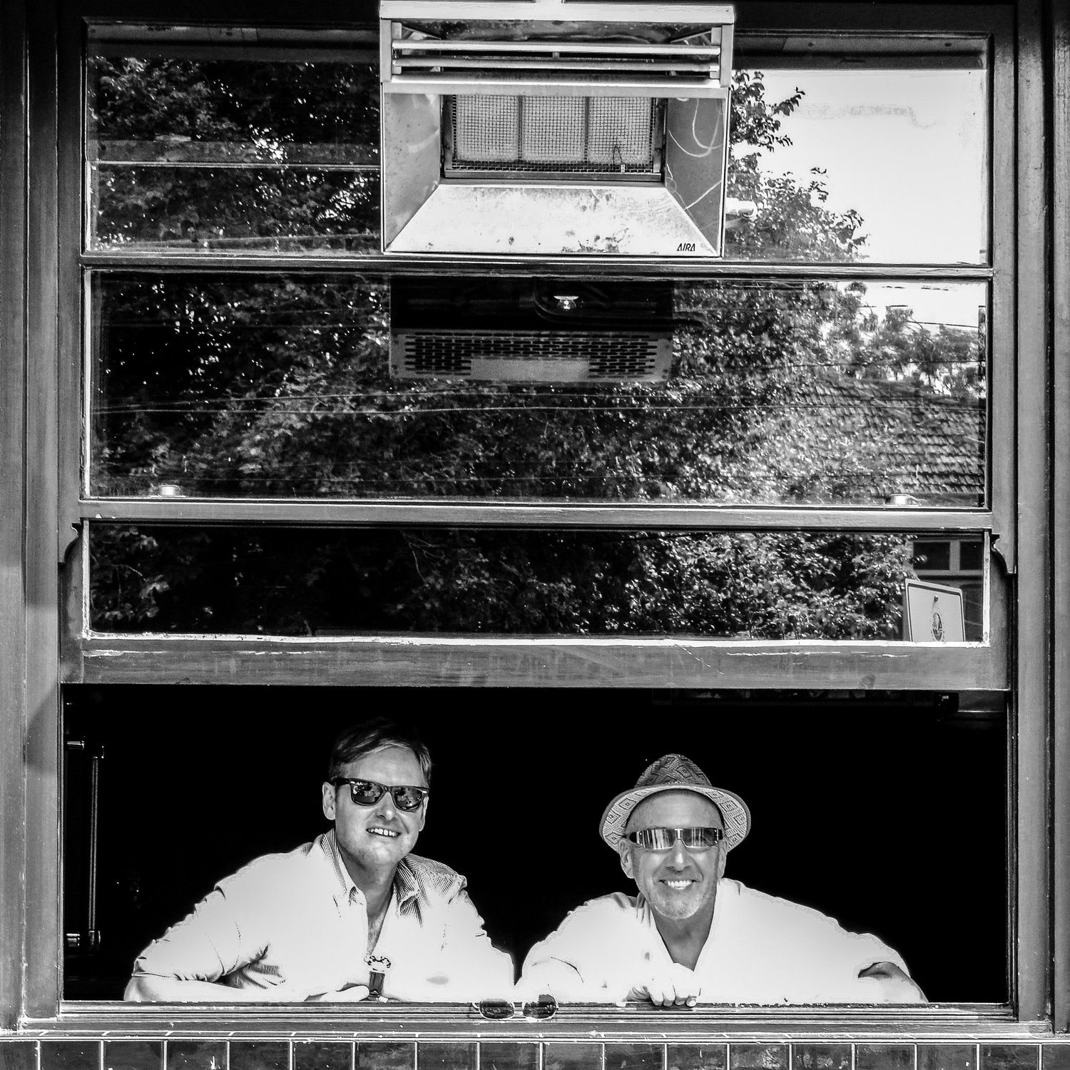 Photo en noir et blanc de deux hommes souriants regardant par une fenêtre ouverte par Jérémie Botte.