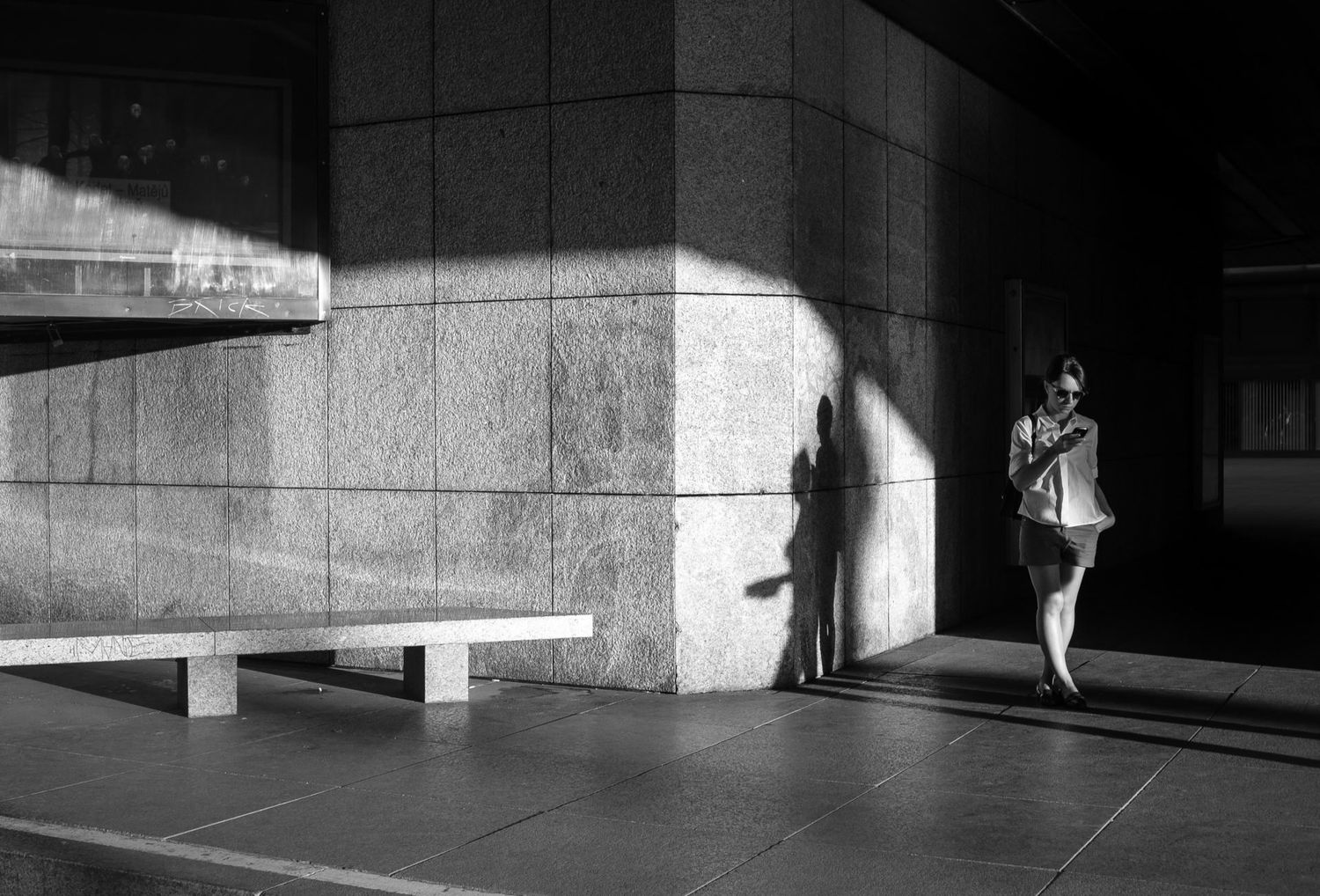 Une femme regarde son téléphone devant un grand bâtiment moderne en pierre, par Jérémie Botte.