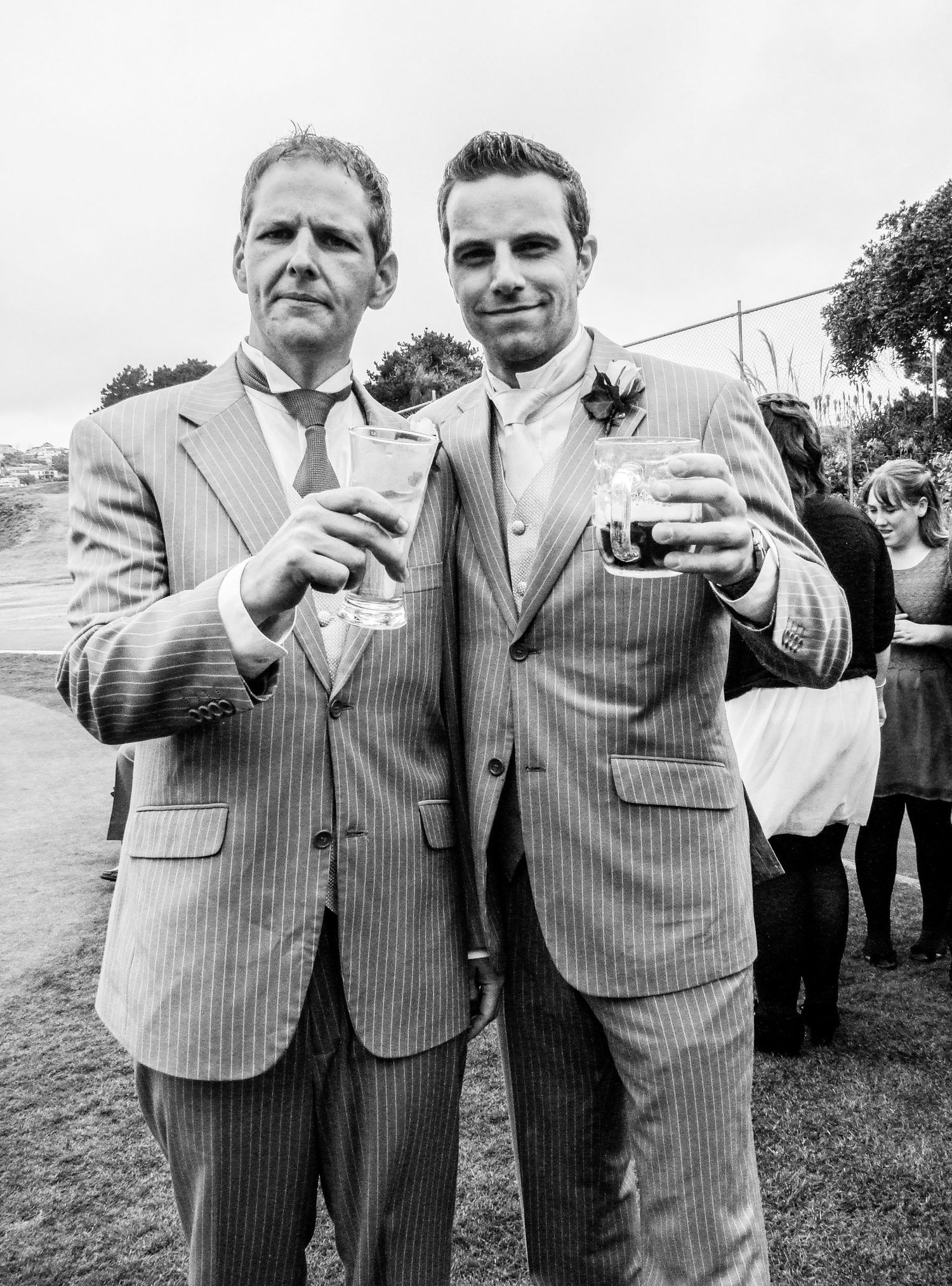Portrait en noir et blanc de deux hommes en costume souriant et trinquant avec leurs verres lors d'un mariage, par Jérémie Botte.