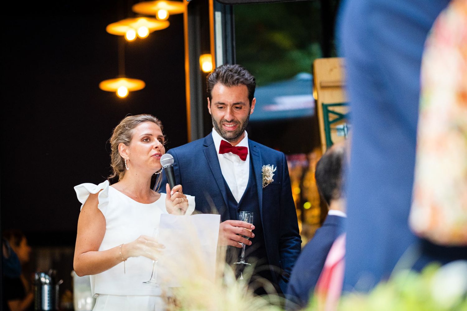 Une mariée s'exprimant au micro aux côtés de son époux en costume lors d'une réception, par Jérémie Botte.