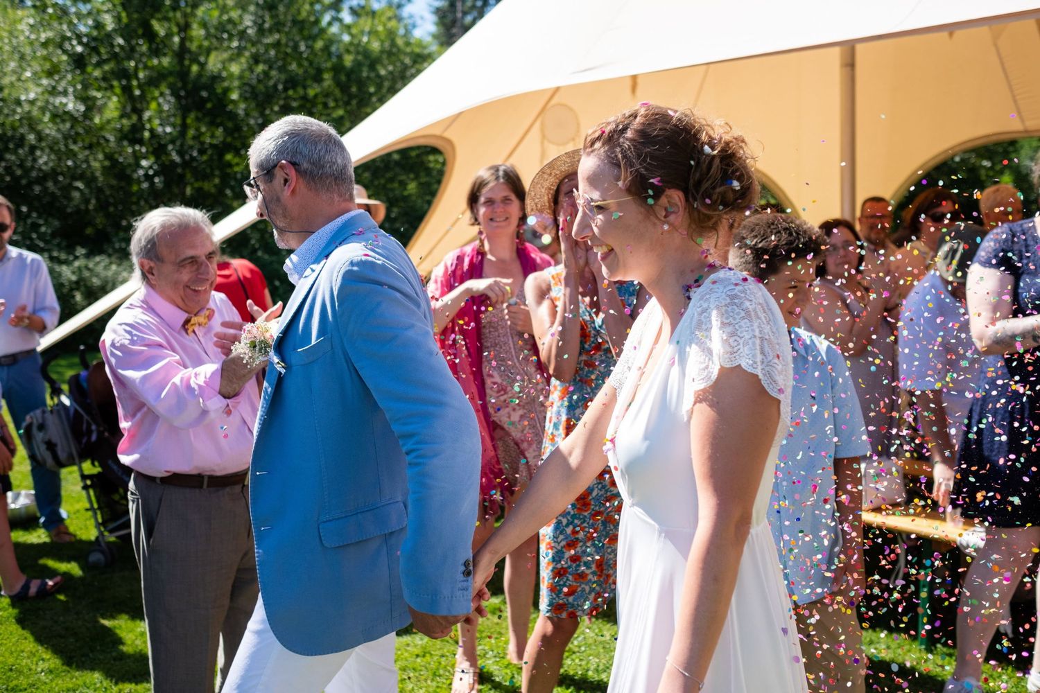 Le couple de mariés avance tout sourire sous une pluie de confettis multicolores au milieu des invités, par Jérémie Botte.