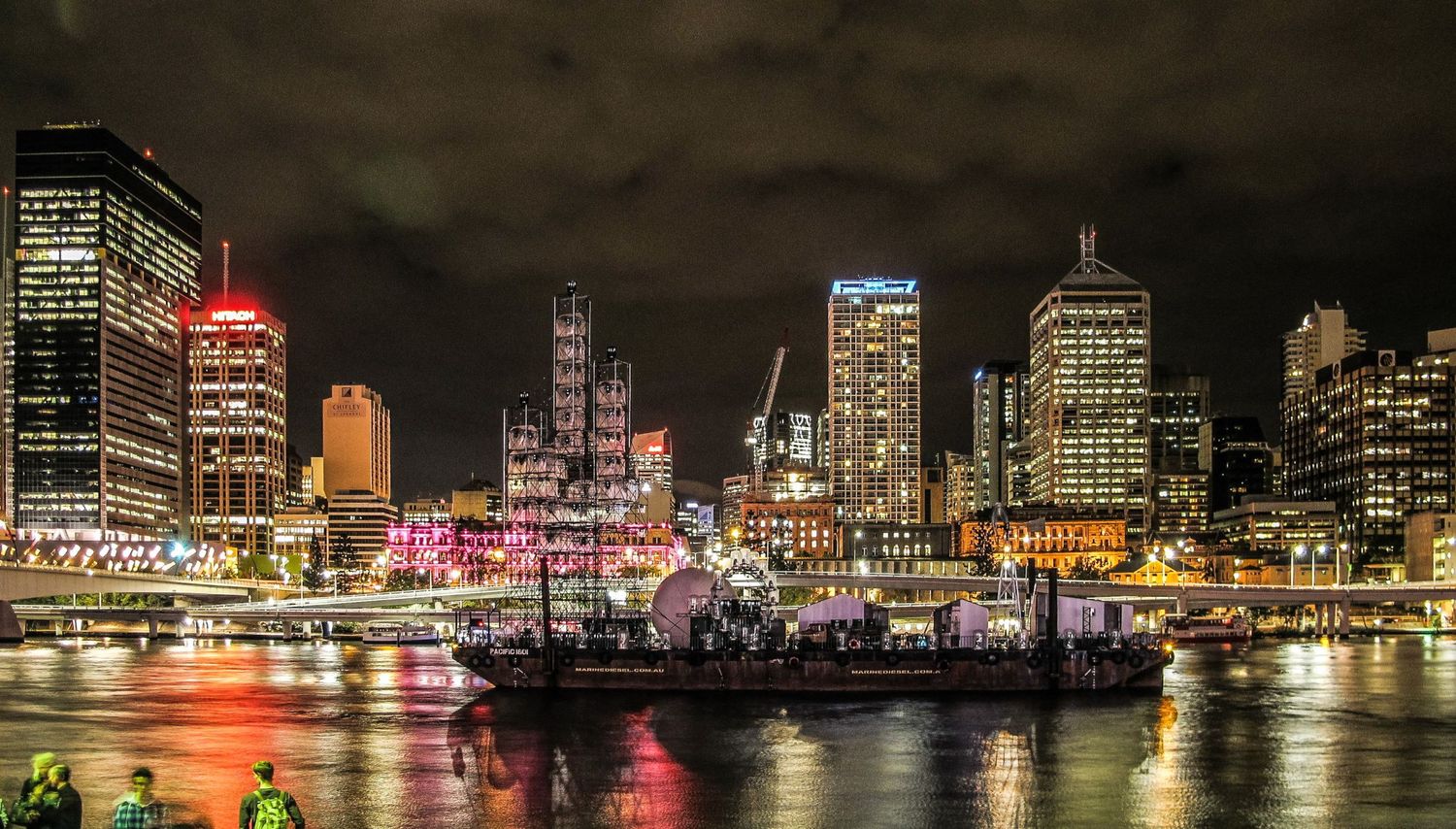 Une vue nocturne de la skyline illuminée de Brisbane qui se reflète sur les eaux du fleuve avec un bateau à quai au premier plan par J Botte