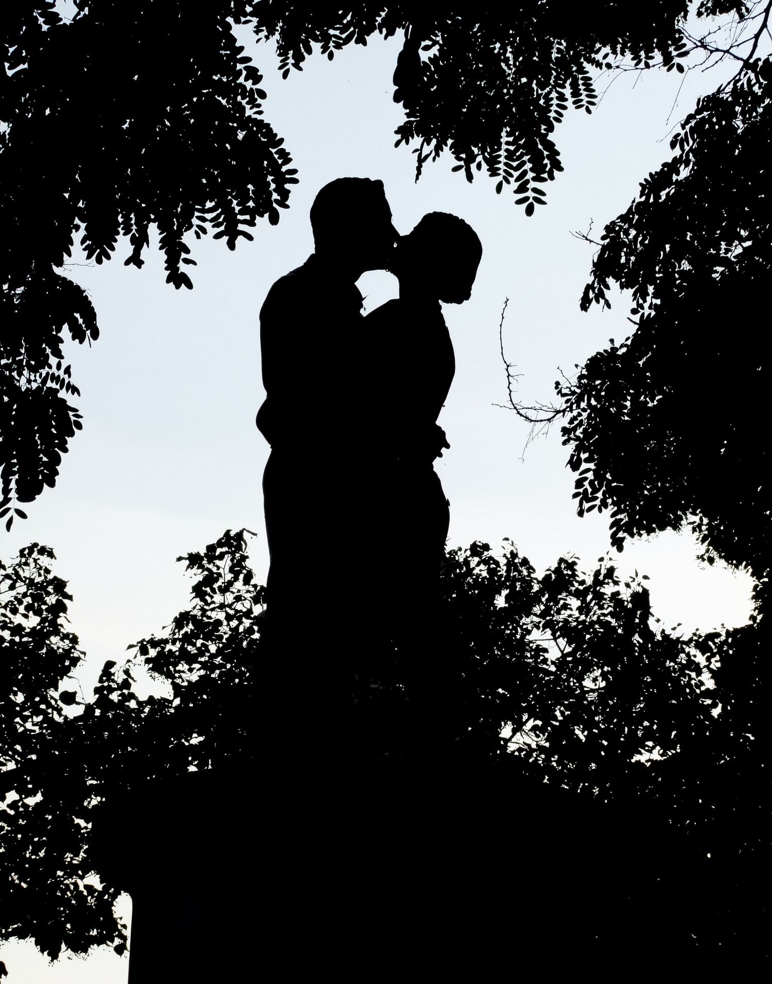 Silhouette en contre-jour d’un couple qui s'embrasse tendrement au milieu des feuillages d'un parc par Jérémie Botte.