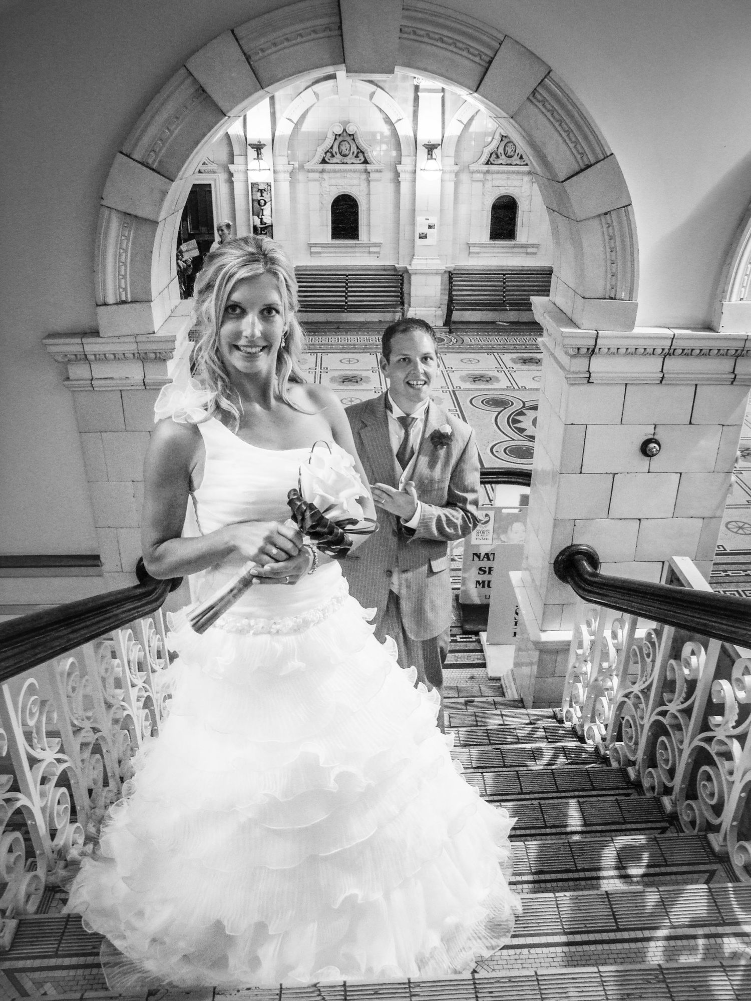Portrait de mariage en noir et blanc, cadré sous une arche en pierre alors que les mariés montent des escaliers, par Jérémie Botte.
