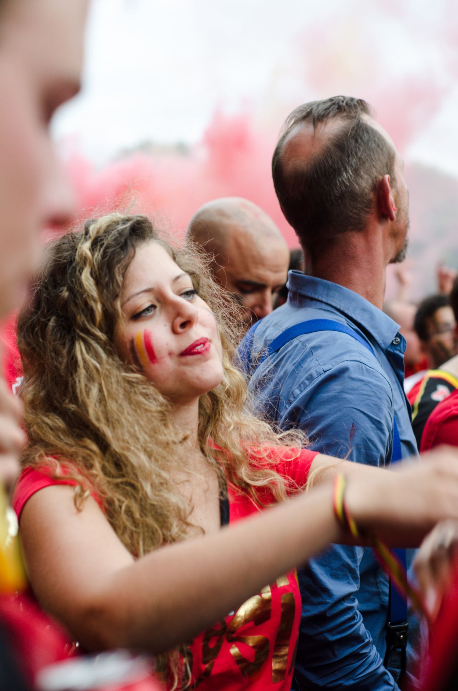 Une supportrice aux cheveux bouclés, maquillée aux couleurs de la Belgique, sourit au milieu d'une foule par Jérémie Botte.