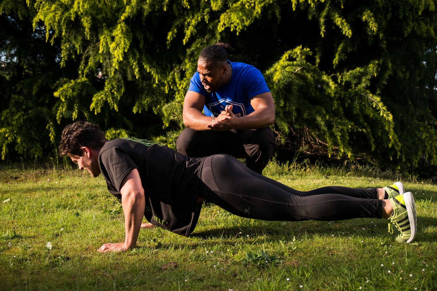 Un homme en pleine séance de gainage sur l'herbe sous le regard attentif de son coach sportif par Jérémie Botte.