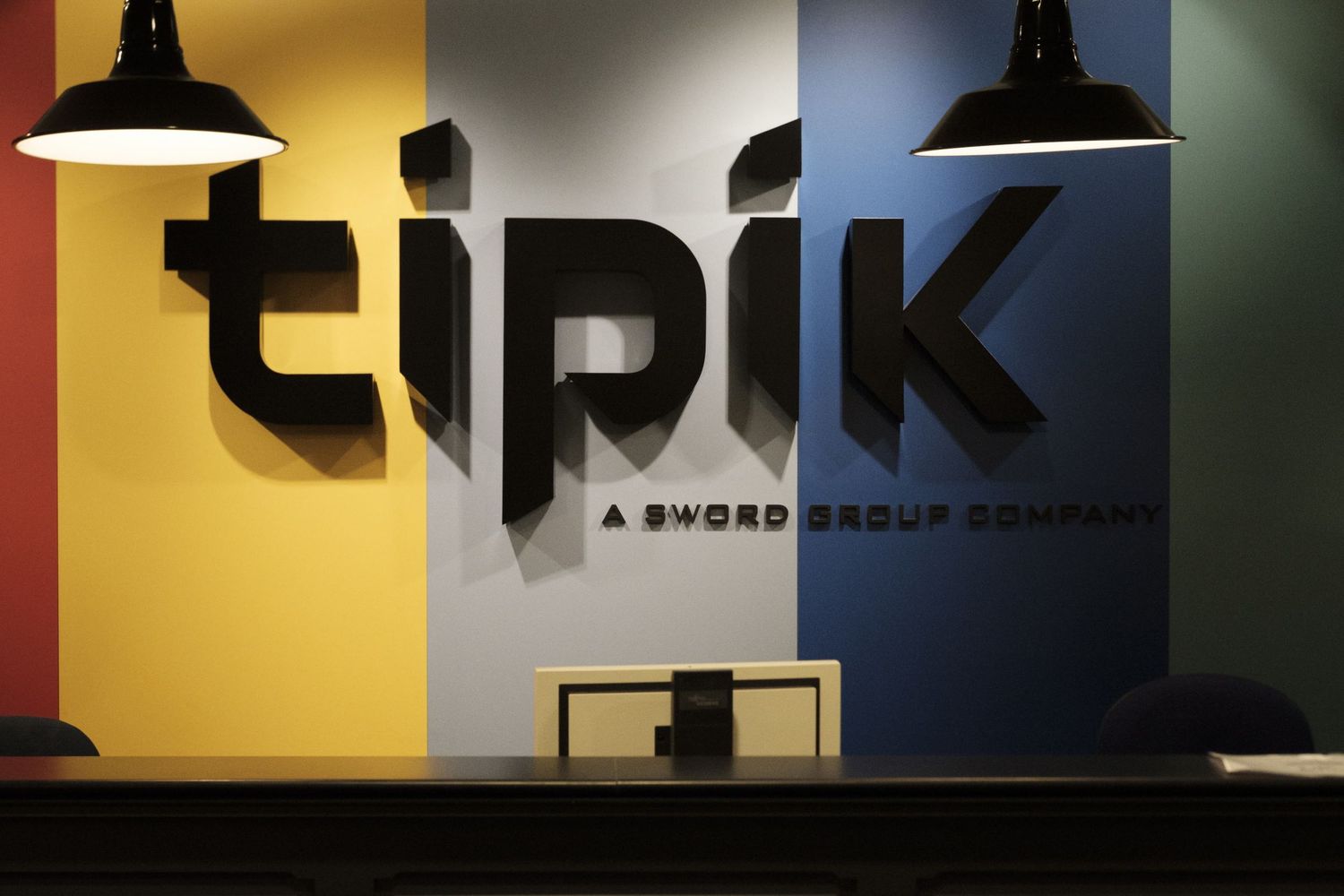 Logo mural noir de l'entreprise Tipik sur un fond coloré rouge, jaune, gris, bleu et vert  sous deux lampes à Bruxelles par Jérémie Botte.