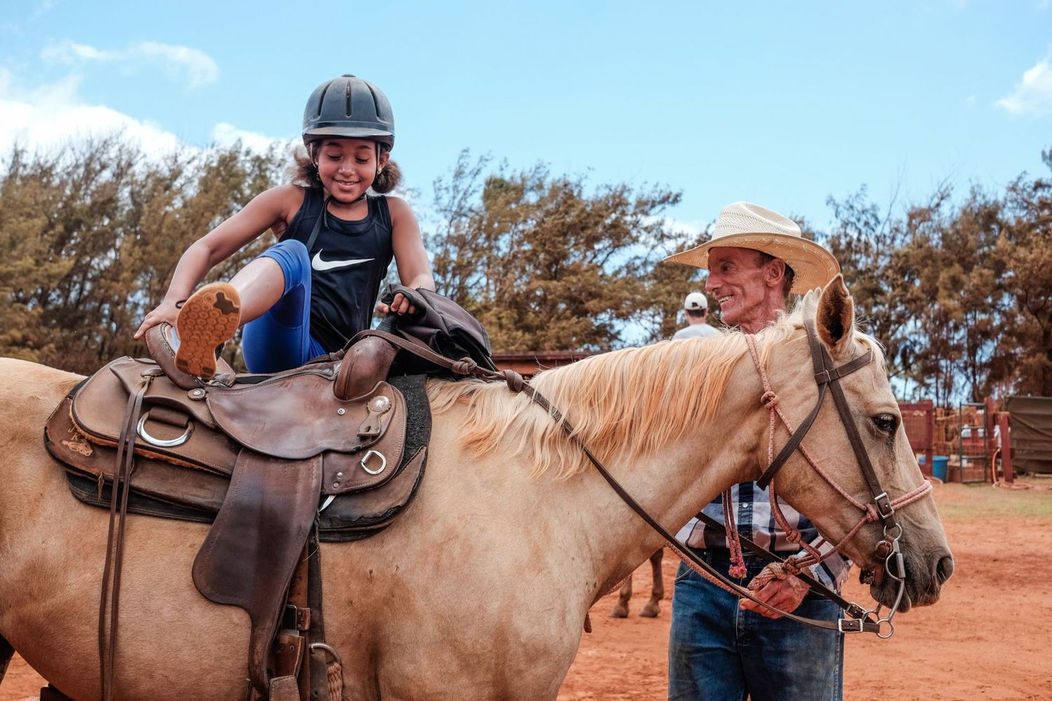 Une jeune fille casquée s'apprête à monter  à cheval tenu par un homme au chapeau de cowboy, par Jérémie Botte.