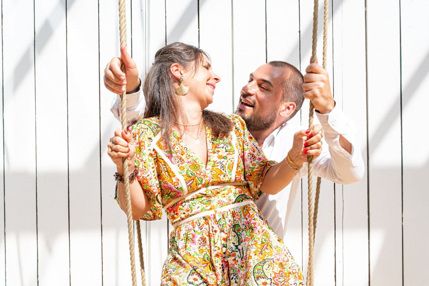 Photographie lumineuse d'un couple souriant s'amusant sur une balançoire en bois devant un mur blanc par le photographe Jérémie Botte.