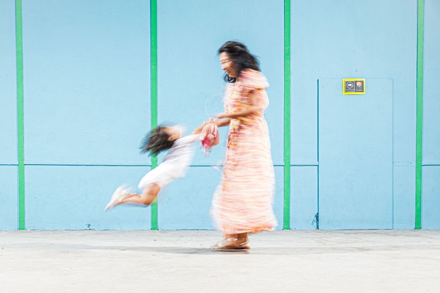 Photographie spontanée d'une mère faisant tourner son enfant en mouvement devant un mur bleu par le photographe Jérémie Botte.