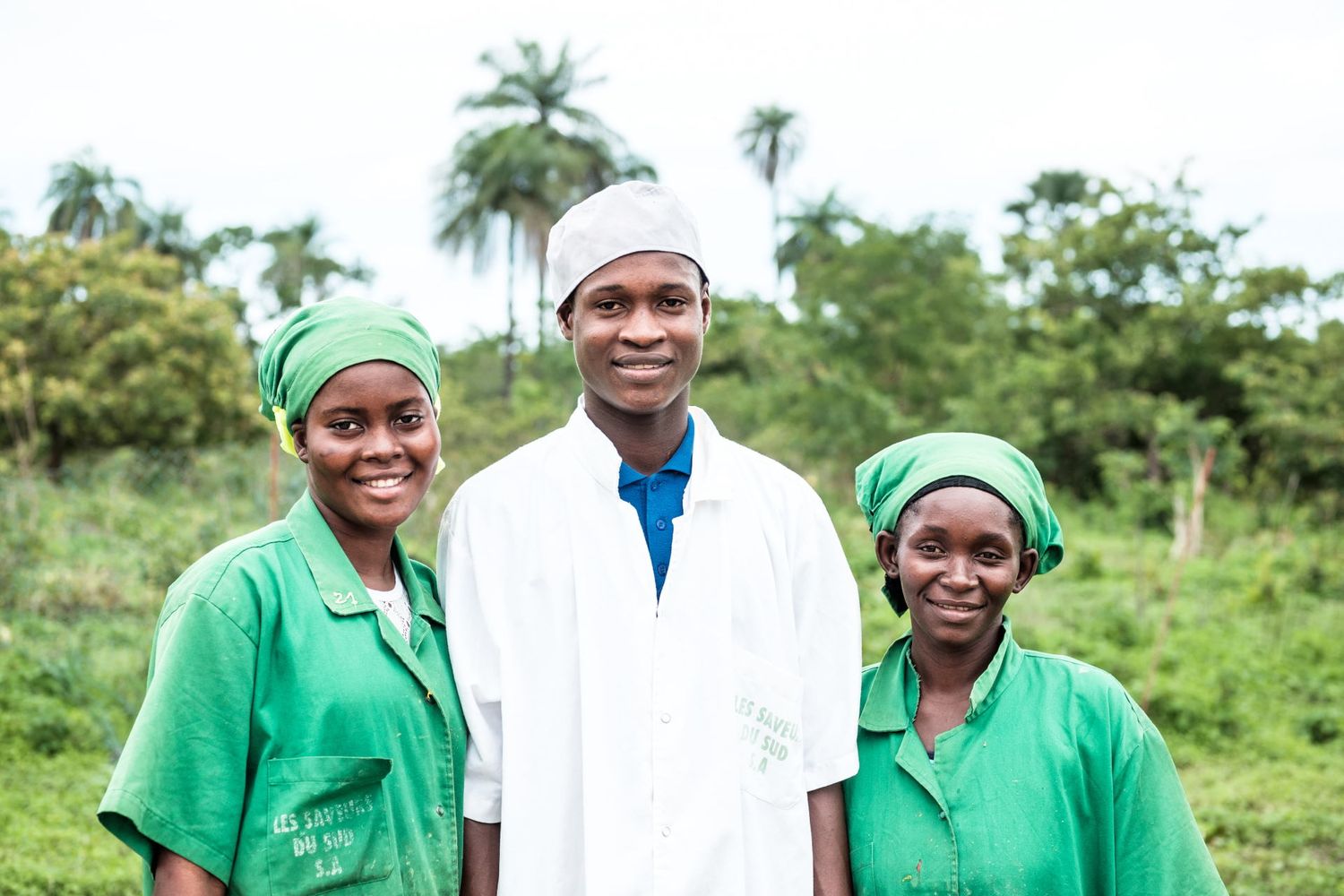 Portrait de trois ouvriers agricoles posant en blouses de travail devant une plantation de palmiers, par Jérémie Botte.