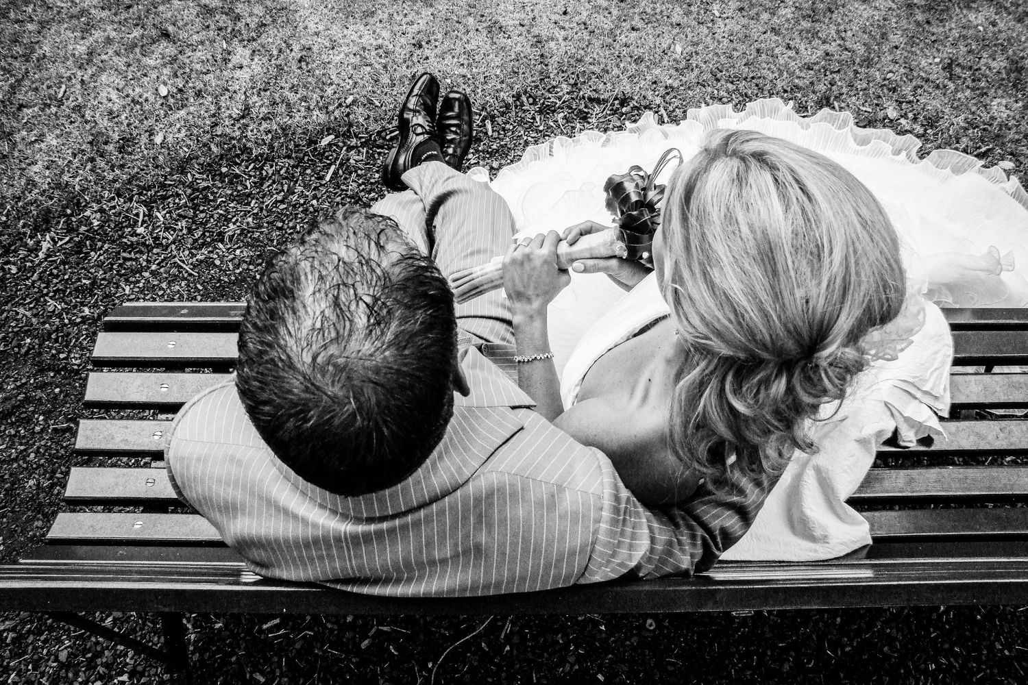 Vue plongeante en noir et blanc sur un couple de mariés assis côte à côte sur un banc en bois, par Jérémie Botte.