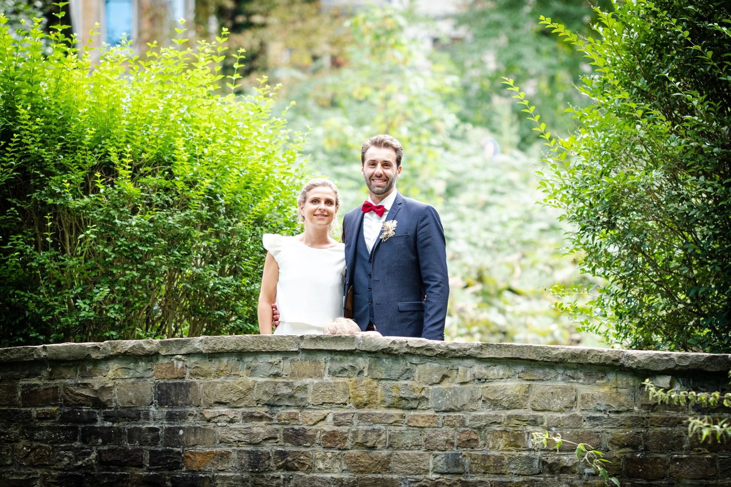 Portrait de mariage du couple de mariés posant avec sourire sur un petit pont en pierre entouré de verdure, par Jérémie Botte.