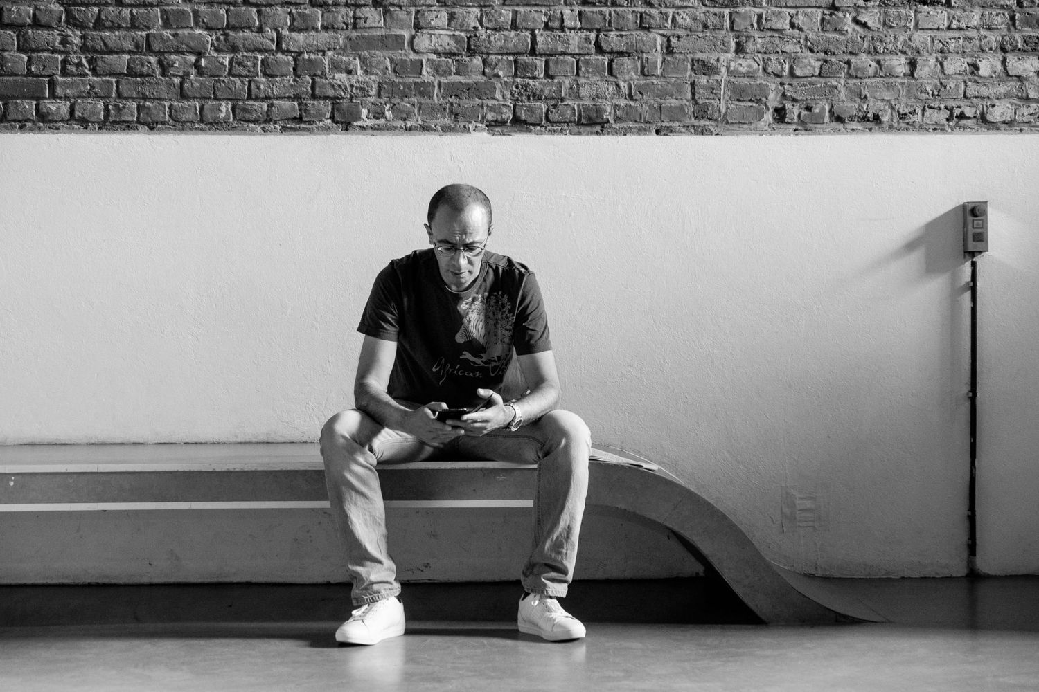 Homme assis sur un banc moderne consultant son smartphone, photo en noir et blanc, par Jérémie Botte.
