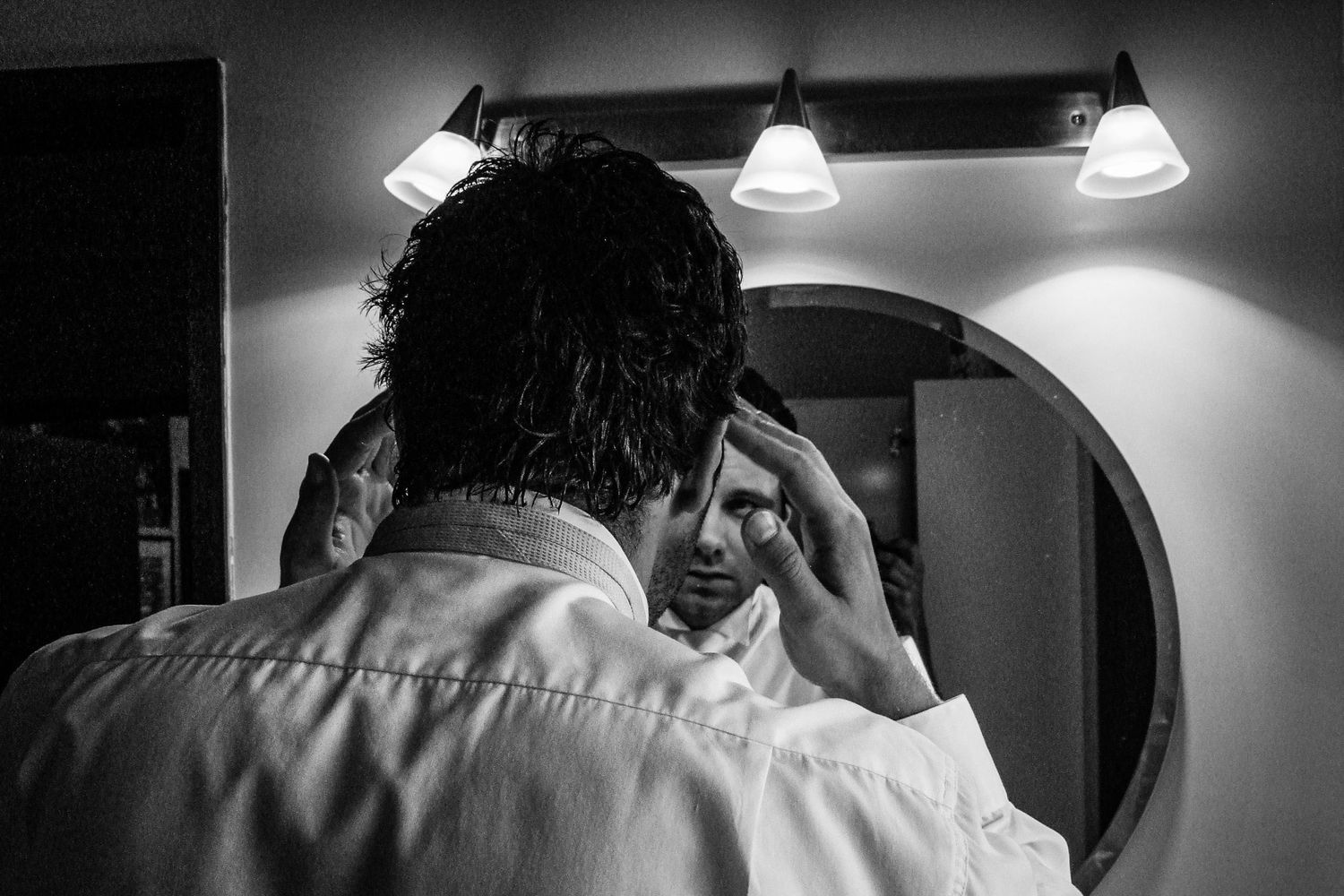 Un homme se préparant devant un miroir de salle de bain, ajustant ses cheveux ou sa cravate, photo noir et blanc par Jérémie Botte.