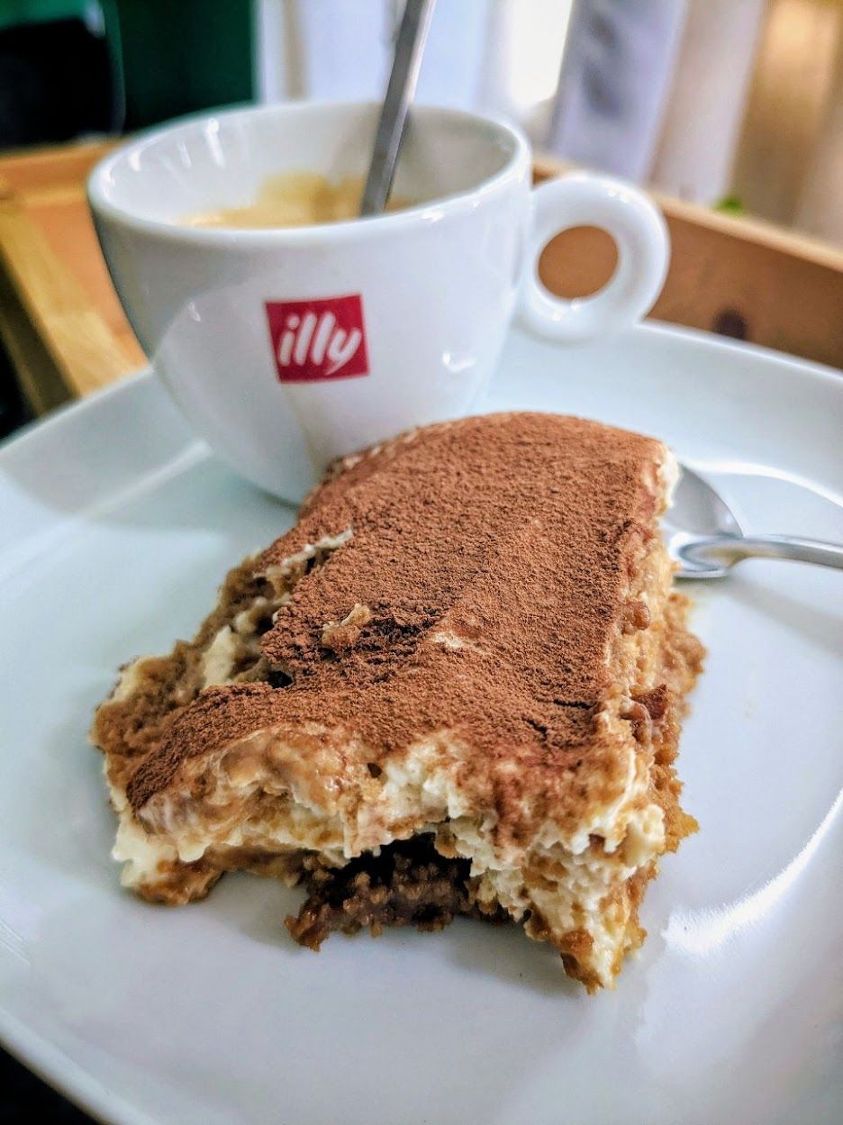 Une part de tiramisu saupoudrée de cacao à côté d'une tasse de café, par Jérémie Botte.