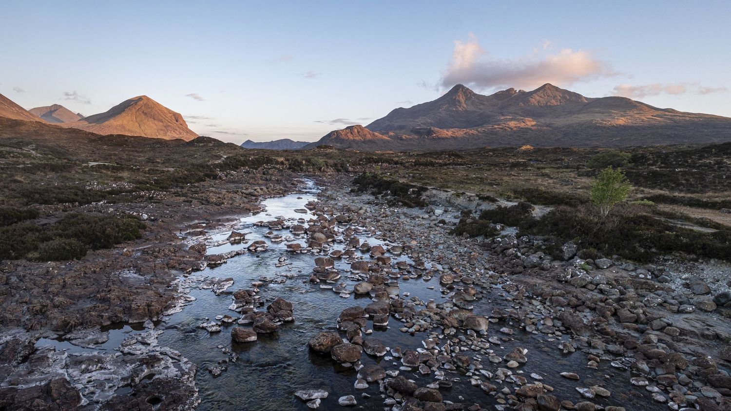 Une rivière parsemée de rochers s'écoule face à de hautes montagnes écossaises au crépuscule par Jérémie Botte.