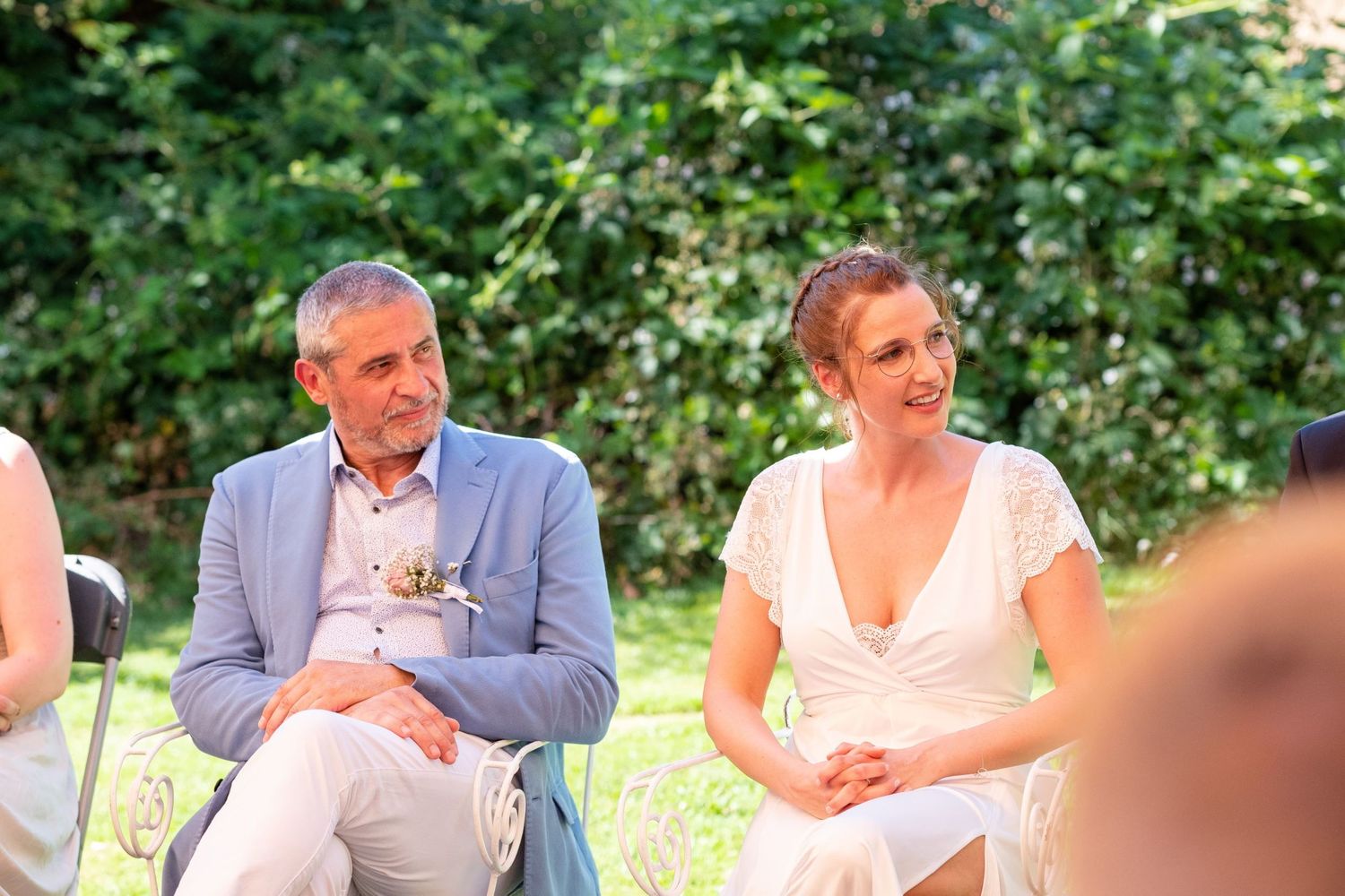 Un homme en veste bleue et une mariée en robe blanche écoutent attentivement un discours assis dans un jardin, par Jérémie Botte.