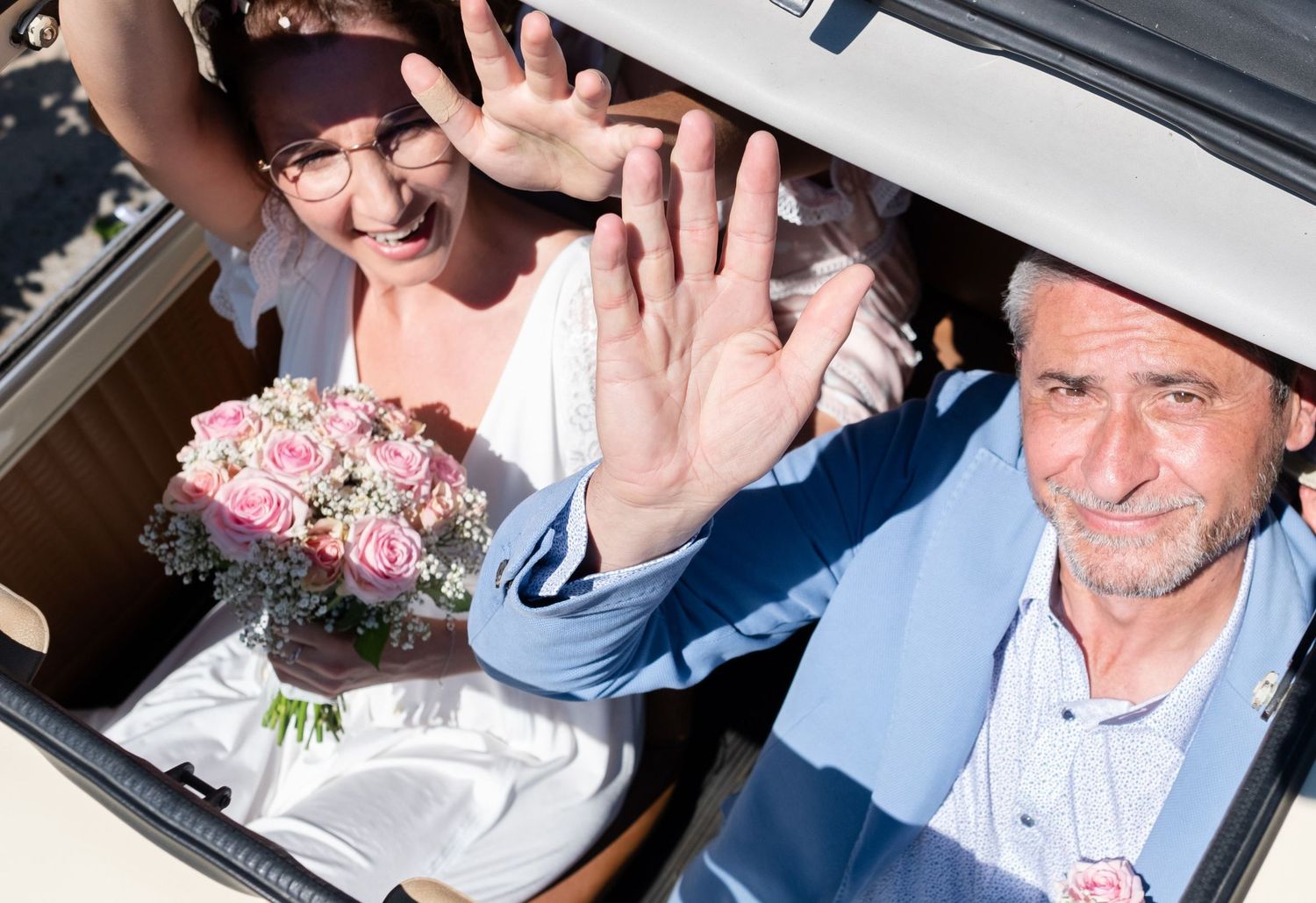 Un couple de mariés tout sourire fait coucou de la main depuis le toit ouvert de leur voiture, par Jérémie Botte.