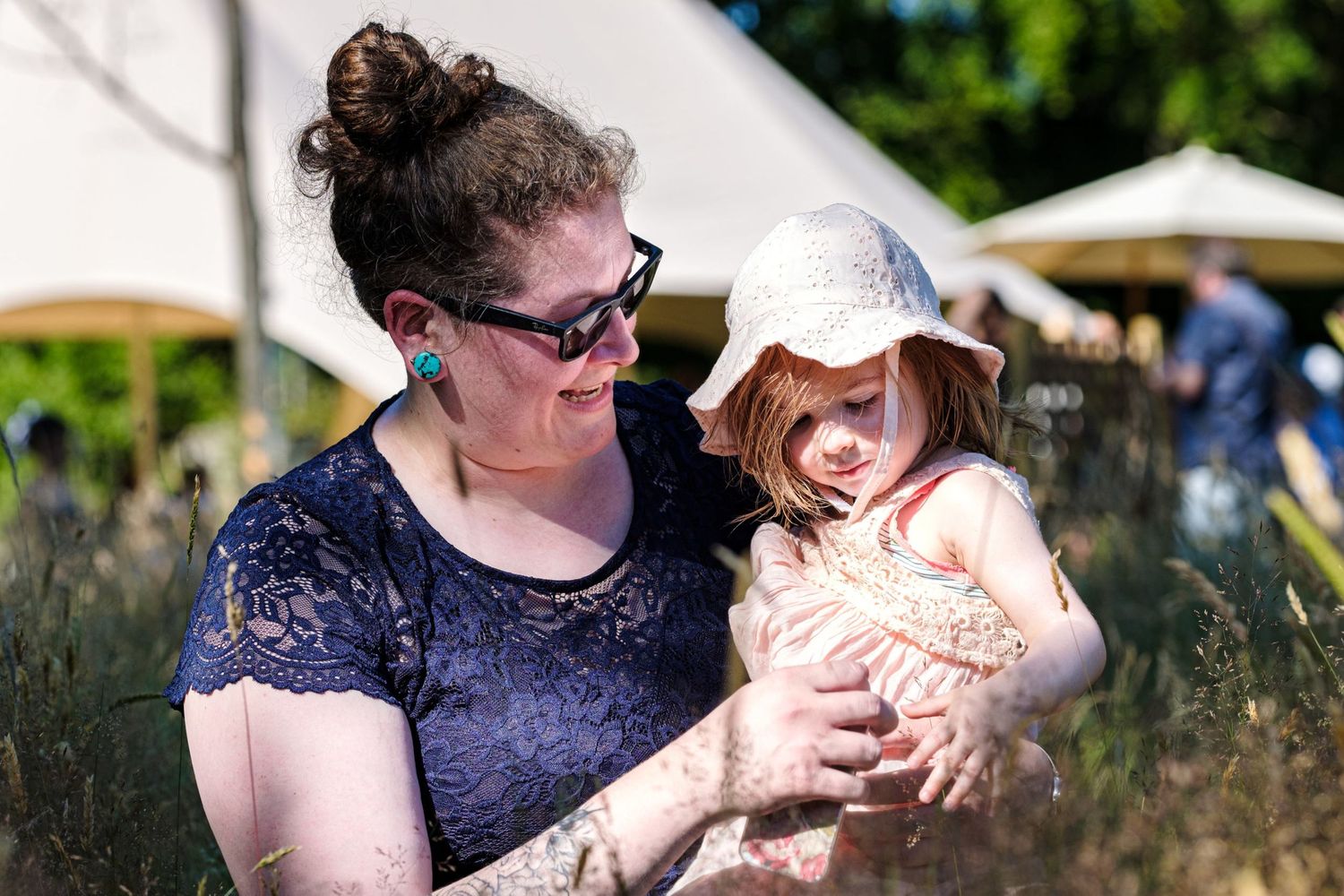 Une mère souriante tient sa petite fille coiffée d'un chapeau au milieu des hautes herbes d'un jardin, par Jérémie Botte.