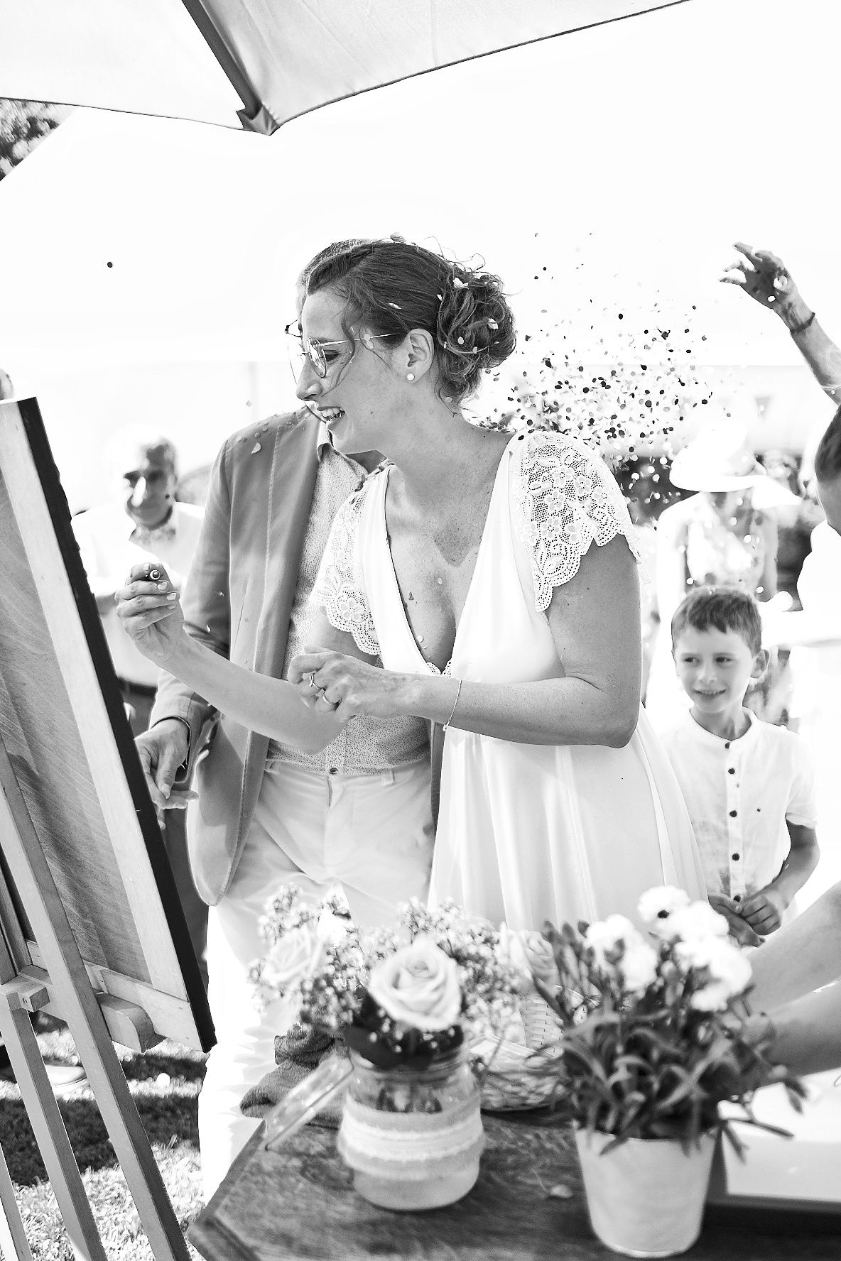 Photographie de mariage spontanée en noir et blanc montrant la mariée souriante en train de peindre sur un chevalet, par Jérémie Botte.