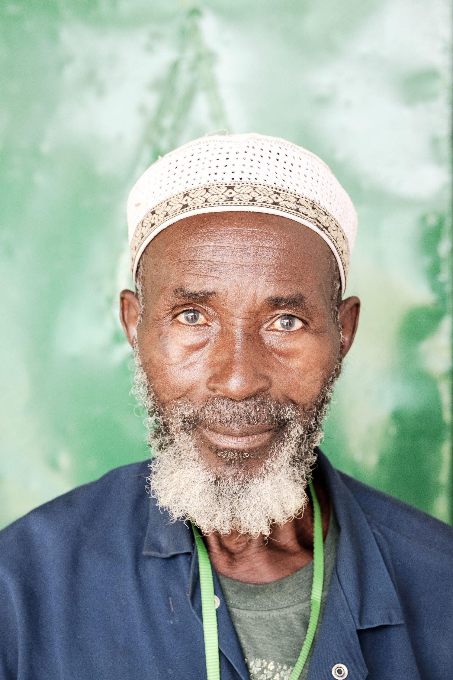 Portrait en gros plan d'un homme âgé barbu portant un bonnet blanc brodé devant un fond vert par Jérémie Botte.