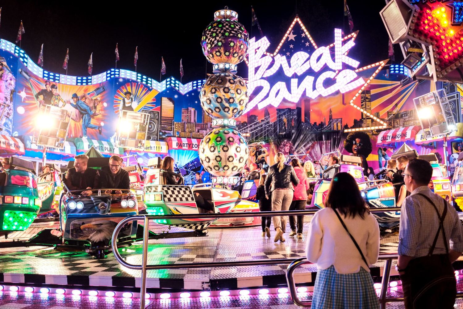 Des passants s'amusent de nuit devant l'enseigne lumineuse rose d'un manège Break Dance par Jérémie Botte.