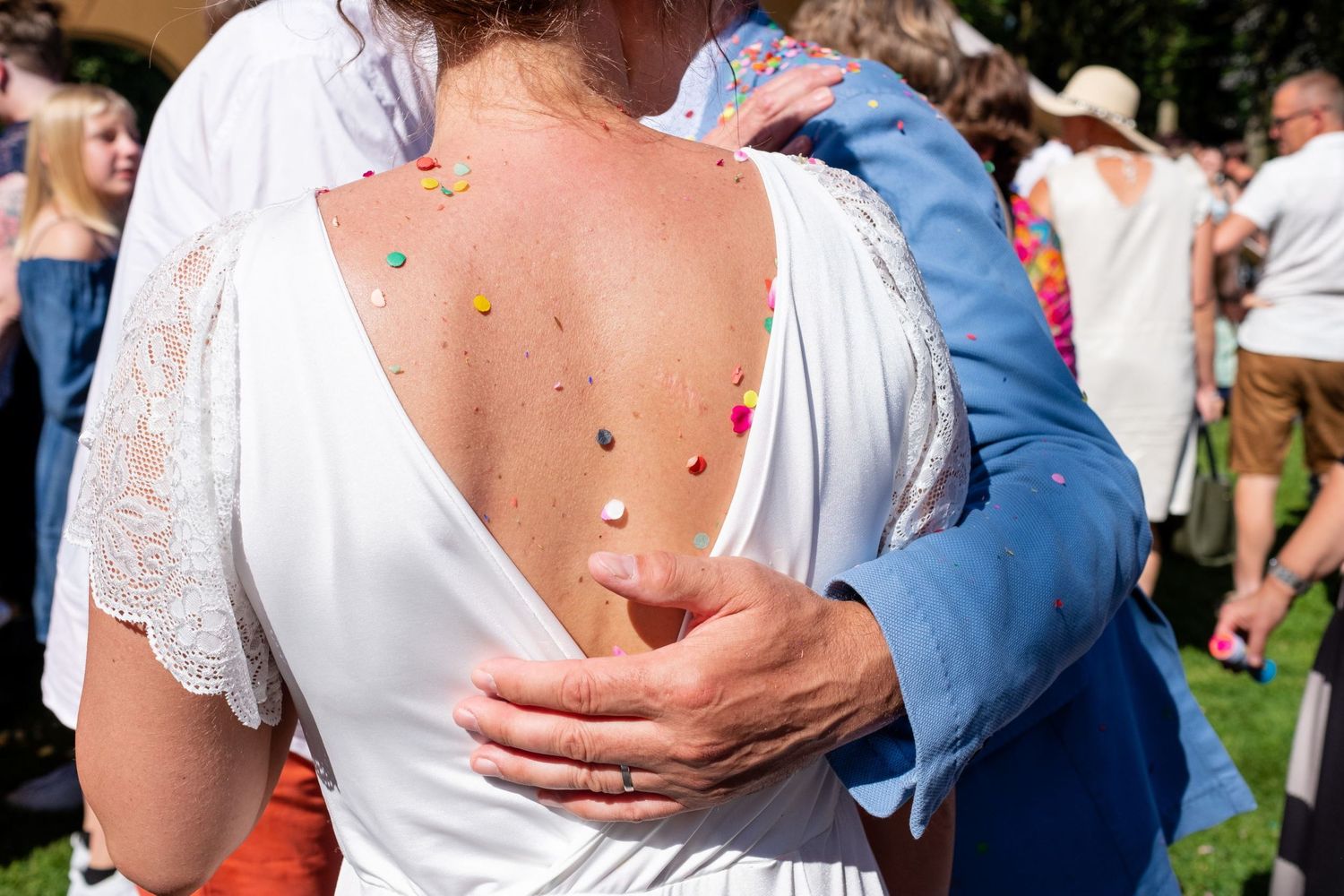 Gros plan sur le dos nu parsemé de confettis d'une mariée tendrement enlacée par son époux en costume bleu, par Jérémie Botte.
