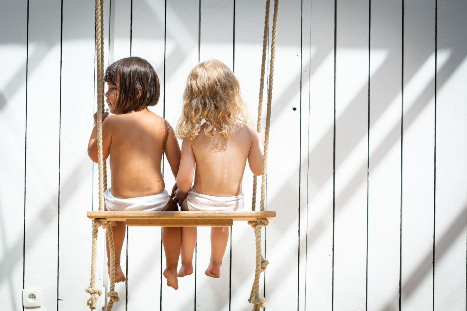Deux jeunes enfants vus de dos assis côte à côte sur une balançoire en bois, par Jérémie Botte.