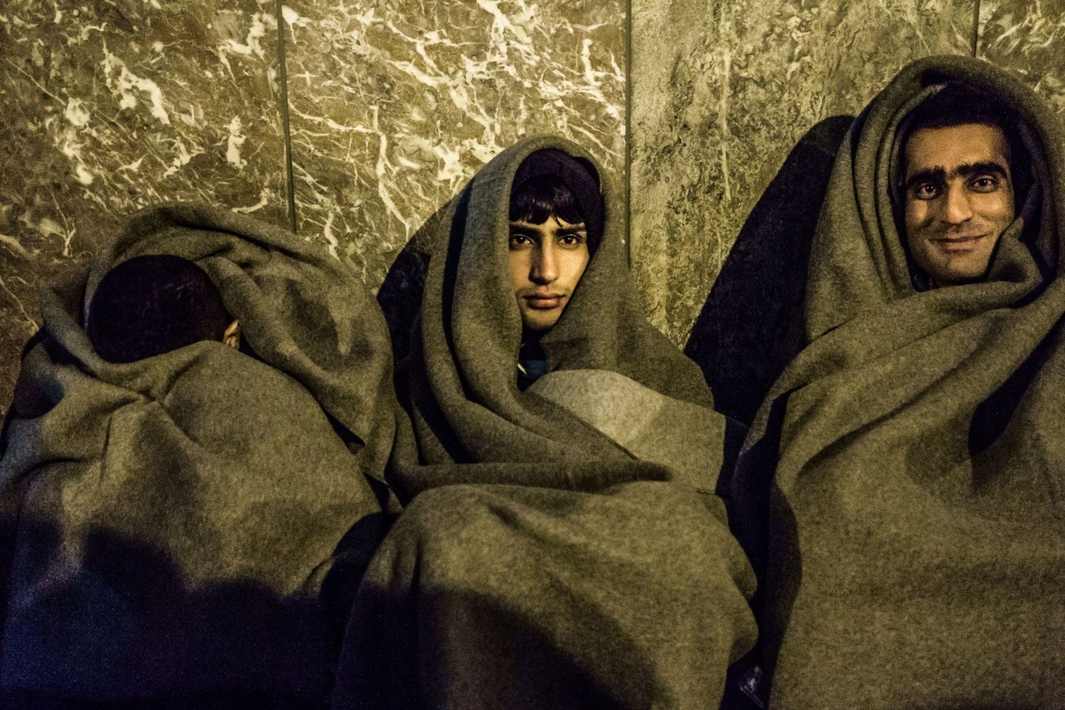 Trois immigrés enveloppées dans de lourdes couvertures grises sont assises contre un mur en marbre par Jérémie Botte.