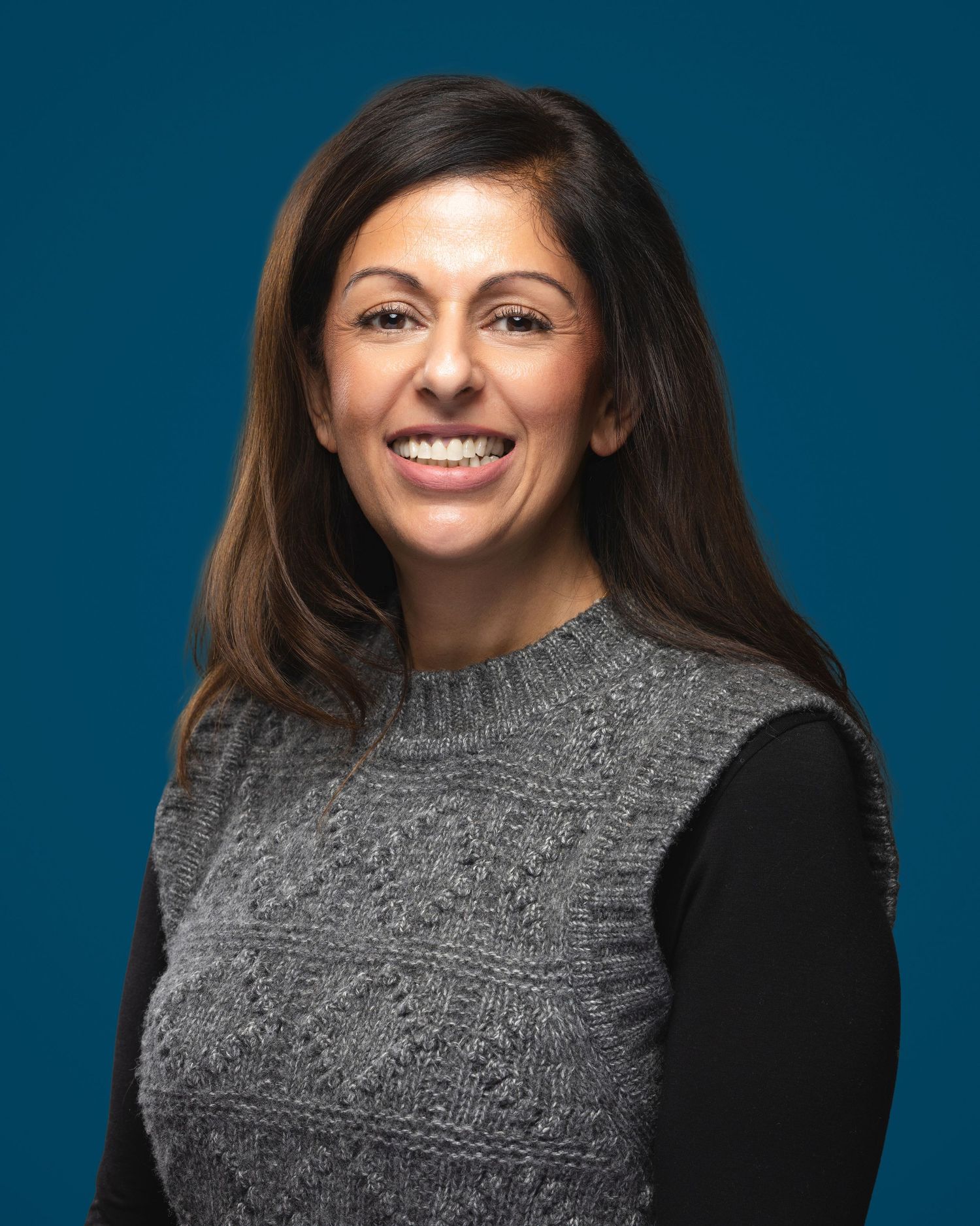 Professional headshot of a smiling woman with dark hair wearing a gray knit vest over a black top on a teal background.