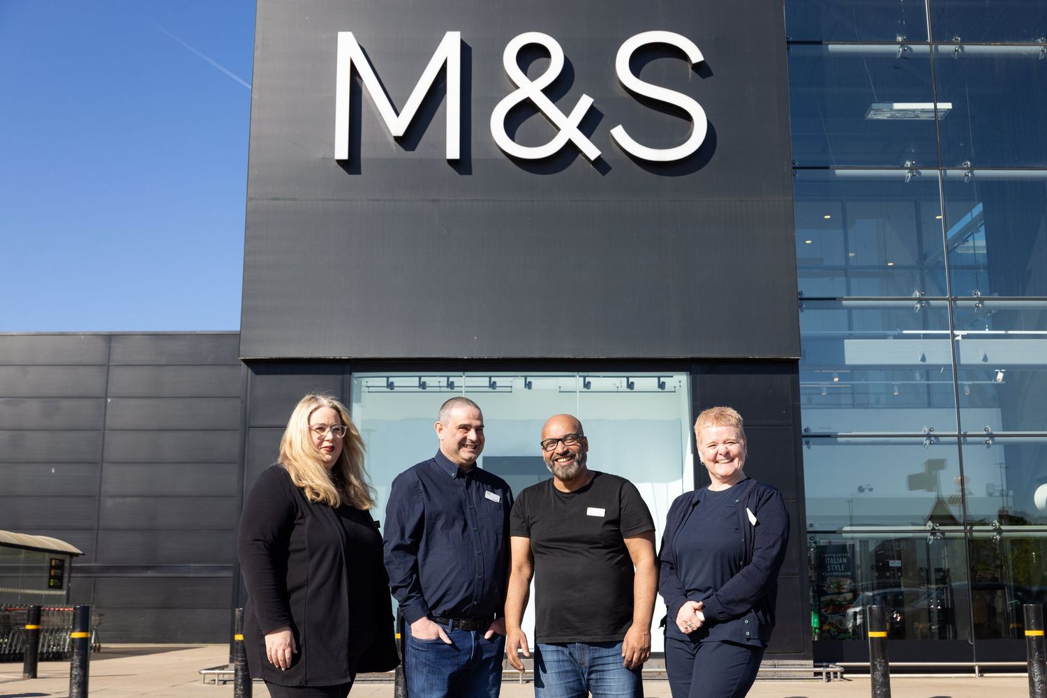 Corporate team photography of four Marks & Spencer employees smiling outside an M&S store entrance on a sunny day.