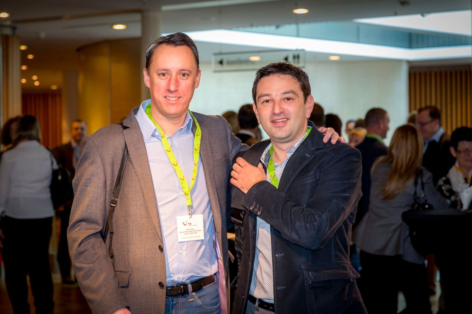 Two smiling men wearing lanyards pose together at a professional networking event with attendees in the background.