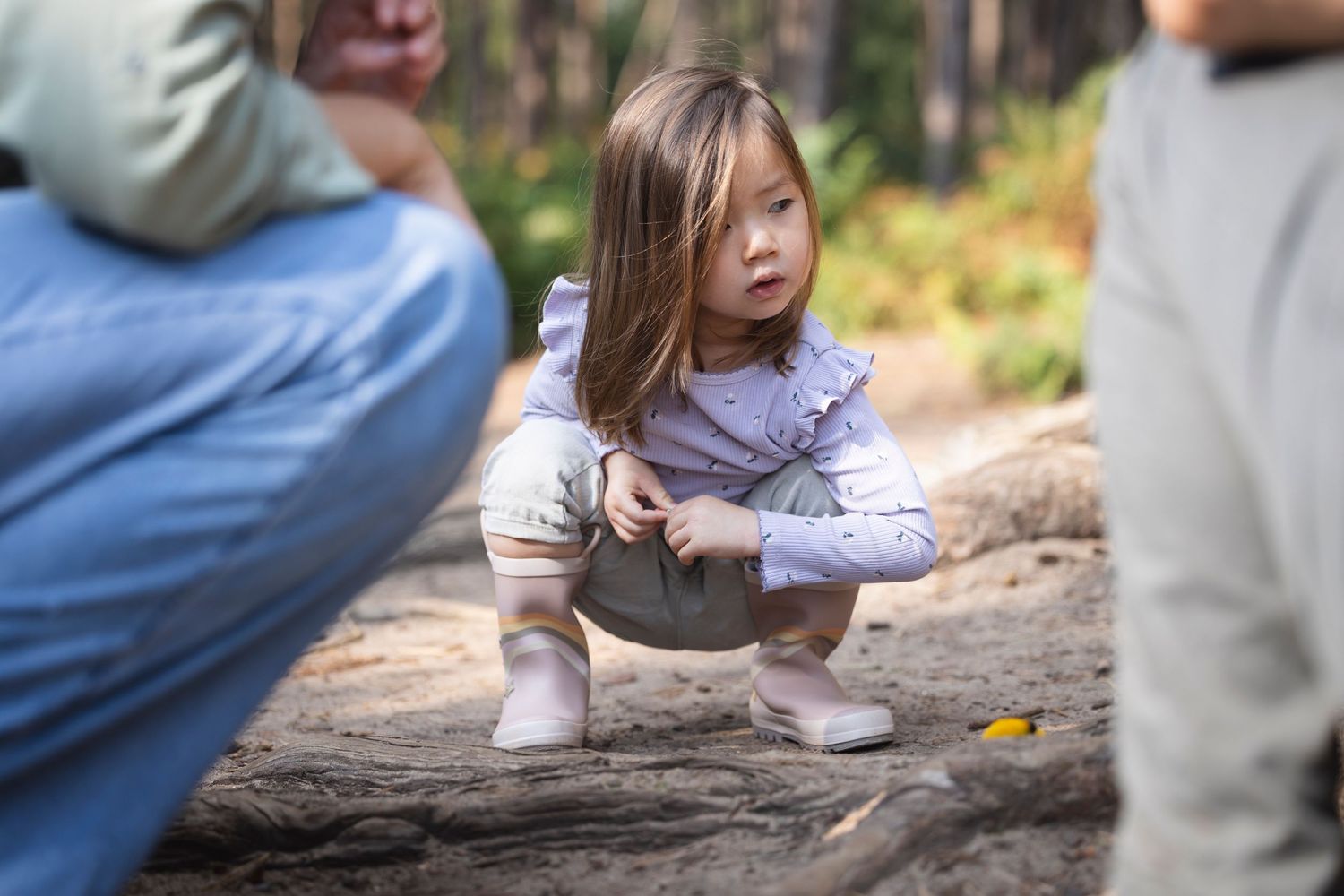 A young toddler girl crouches outdoors in a forest, exploring the ground with curiosity while adults stand nearby.