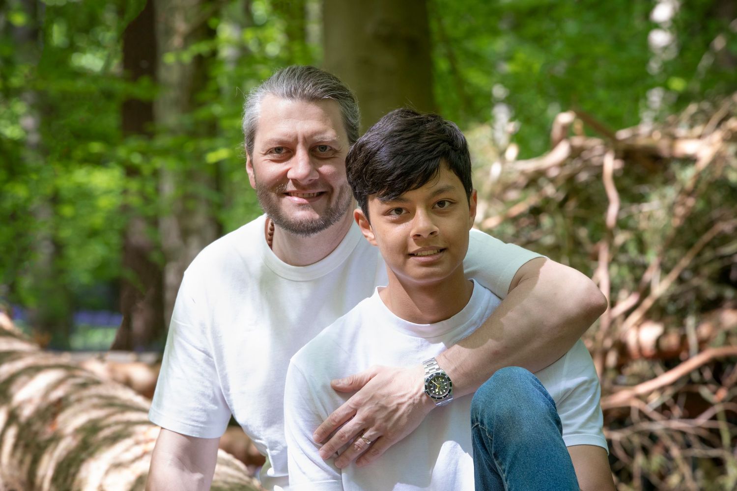 A smiling man and boy in white t-shirts pose together among fallen logs in a lush green forest.