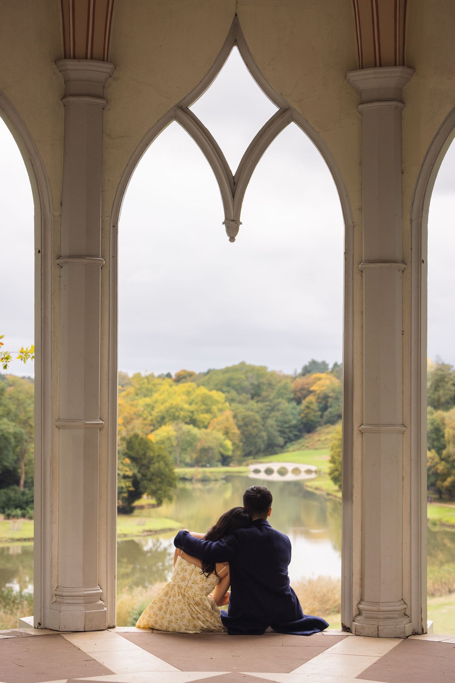 Couple sitting together under Gothic arches overlooking a serene lake and autumn trees in a romantic outdoor setting.