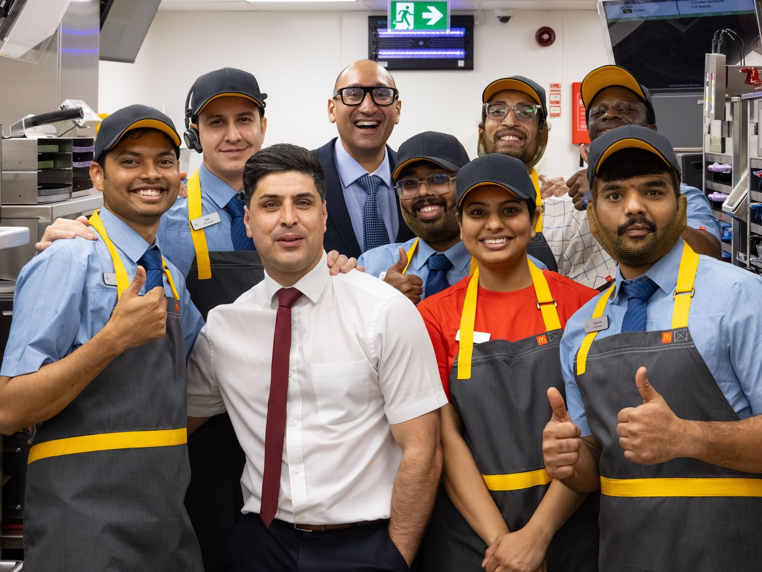 Group of smiling McDonald's employees in uniform giving thumbs up with manager in restaurant kitchen.