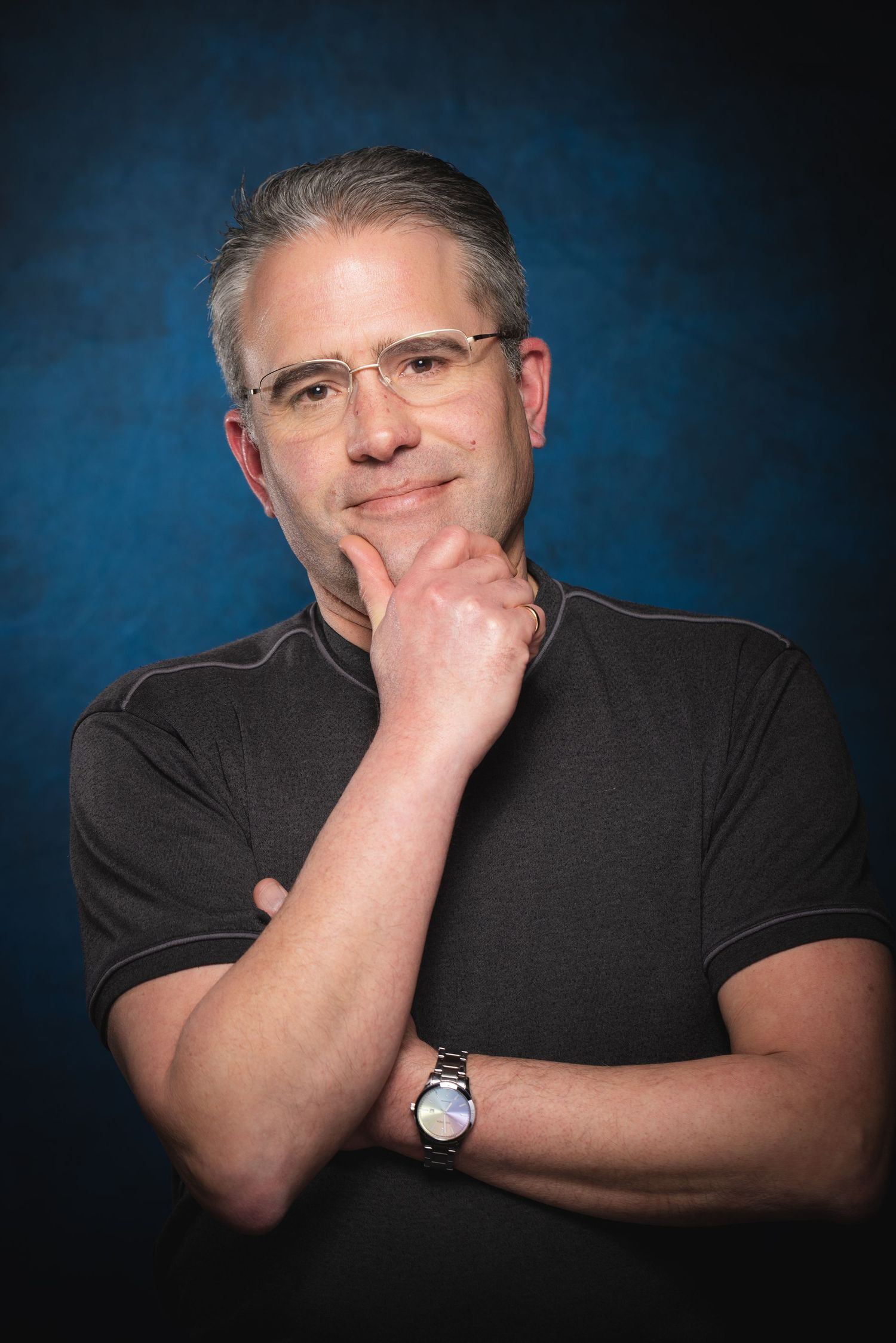 Middle-aged man with gray hair and glasses poses thoughtfully with hand on chin against blue background.
