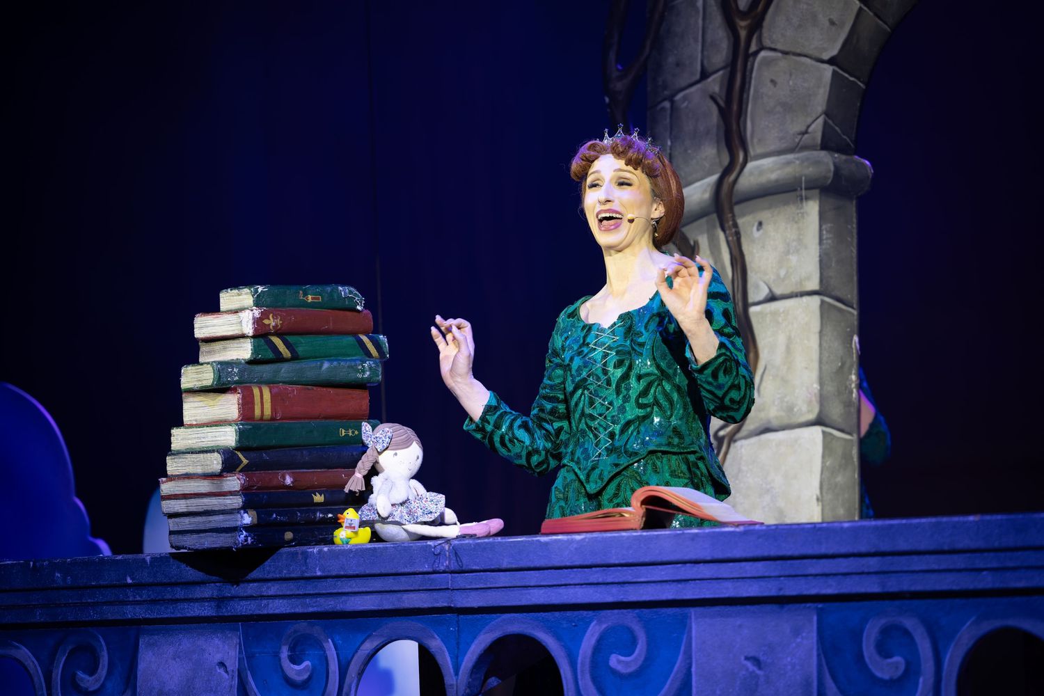 Princess Fiona with floral crown performs beside stacked books on stage during Shrek The Musical at Castle Theatre Wellingborough