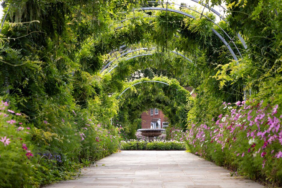 A garden pathway with curved floral archways leads to a red structure, surrounded by lush greenery and pink flowers.