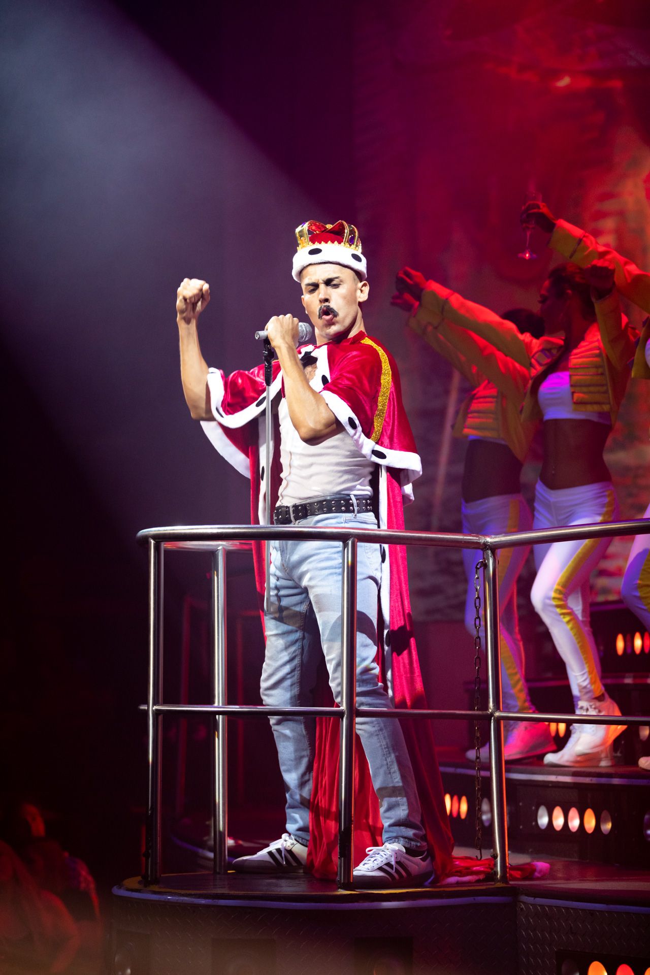 Young performer in regal red cape and crown sings energetically on elevated platform during theatrical stage show.