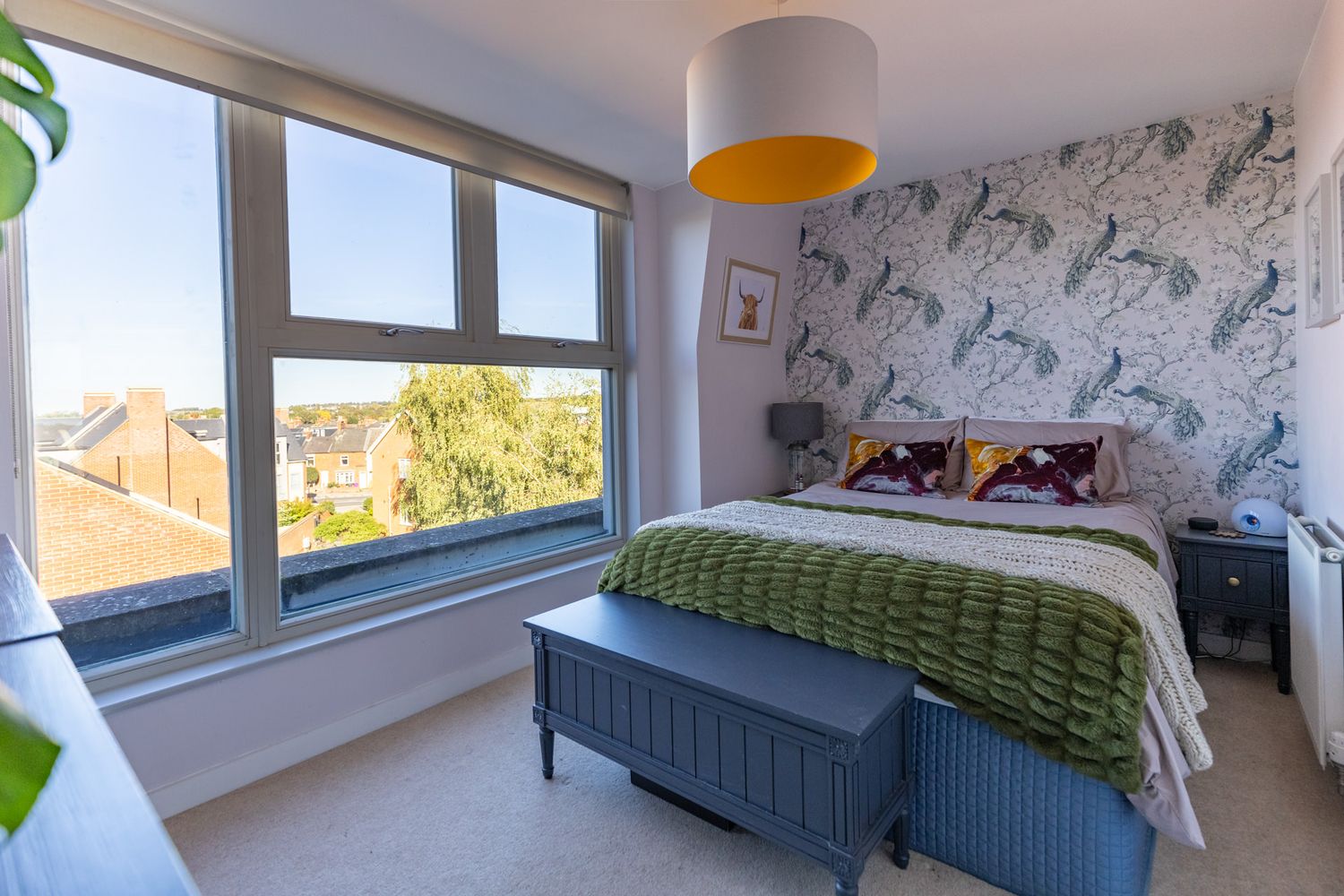 Real estate photography of a bright bedroom with zebra print wallpaper, a large window, a green throw blanket, and a grey bench.