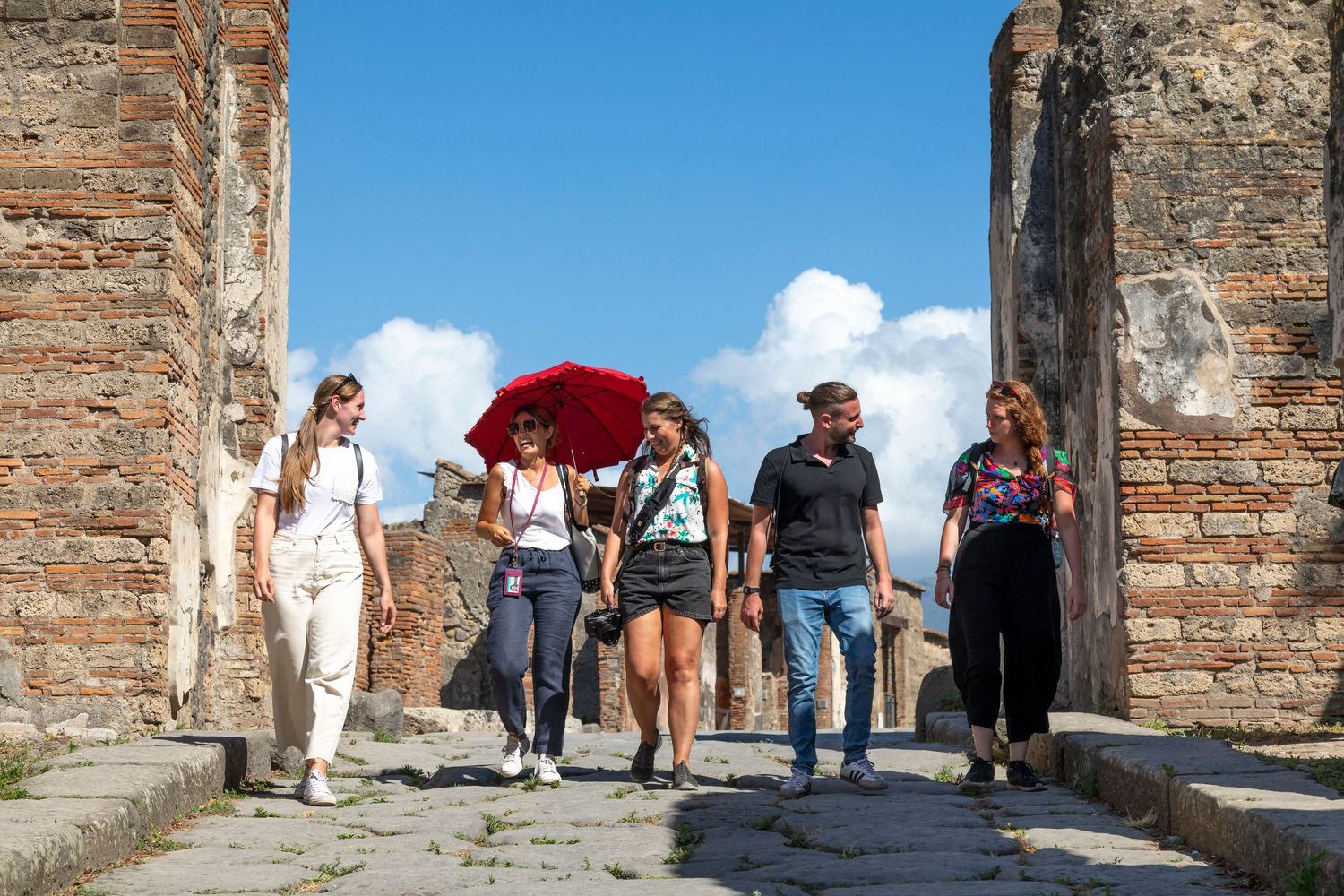 Group of tourists walking along a stone path between ancient brick ruins, one holding a red umbrella, under a blue sky with clouds.