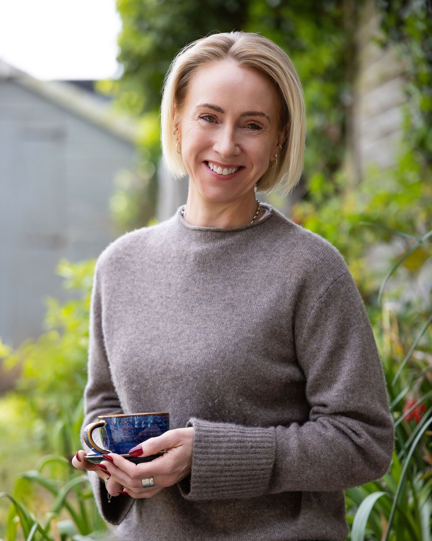 Smiling blonde woman in gray sweater holding blue ceramic mug outdoors in lush green garden setting.