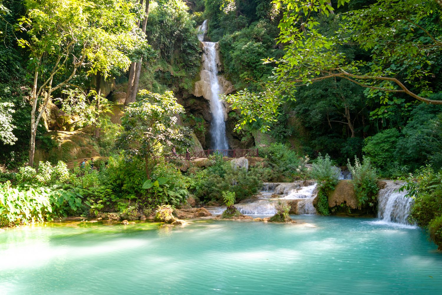 Stunning turquoise waterfall cascades into a crystal-clear pool surrounded by lush green forest in a tropical paradise.