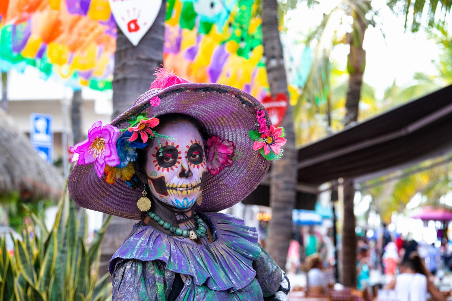 Traditional Day of the Dead figure wearing a purple hat and skeleton makeup at a festive outdoor celebration.