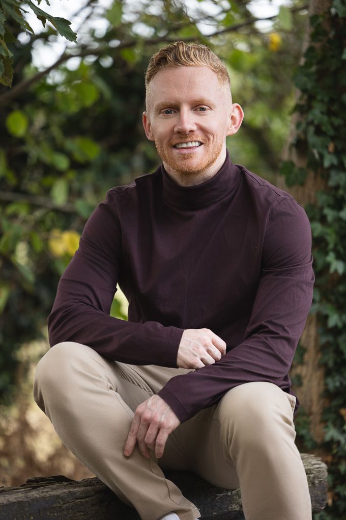 A person in a burgundy turtleneck and khaki pants sits casually outdoors in a garden setting.