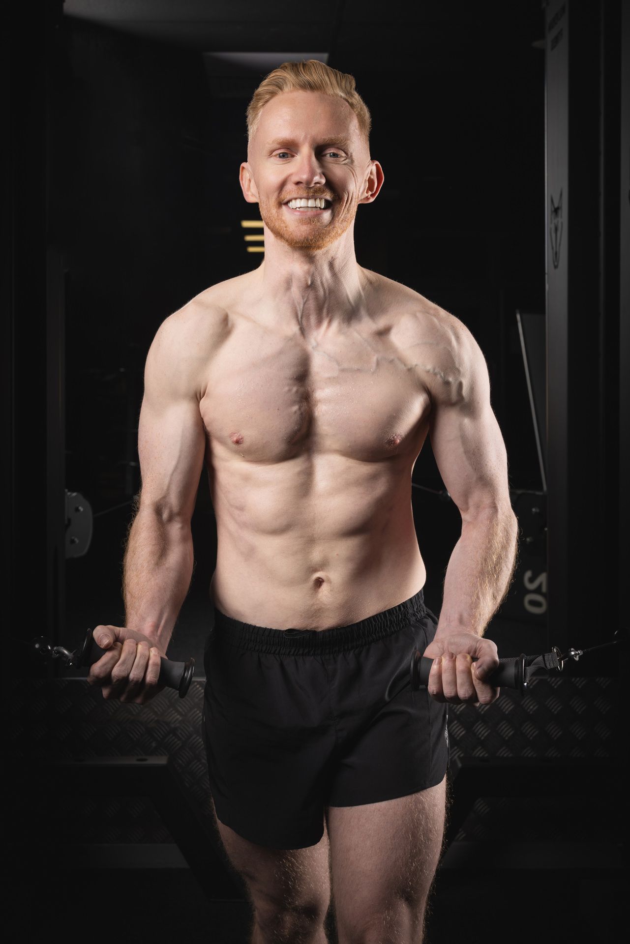 A sports photo shoot. An athletic shirtless person with a muscular physique holding dumbbells in a Hertfordshire Gym.